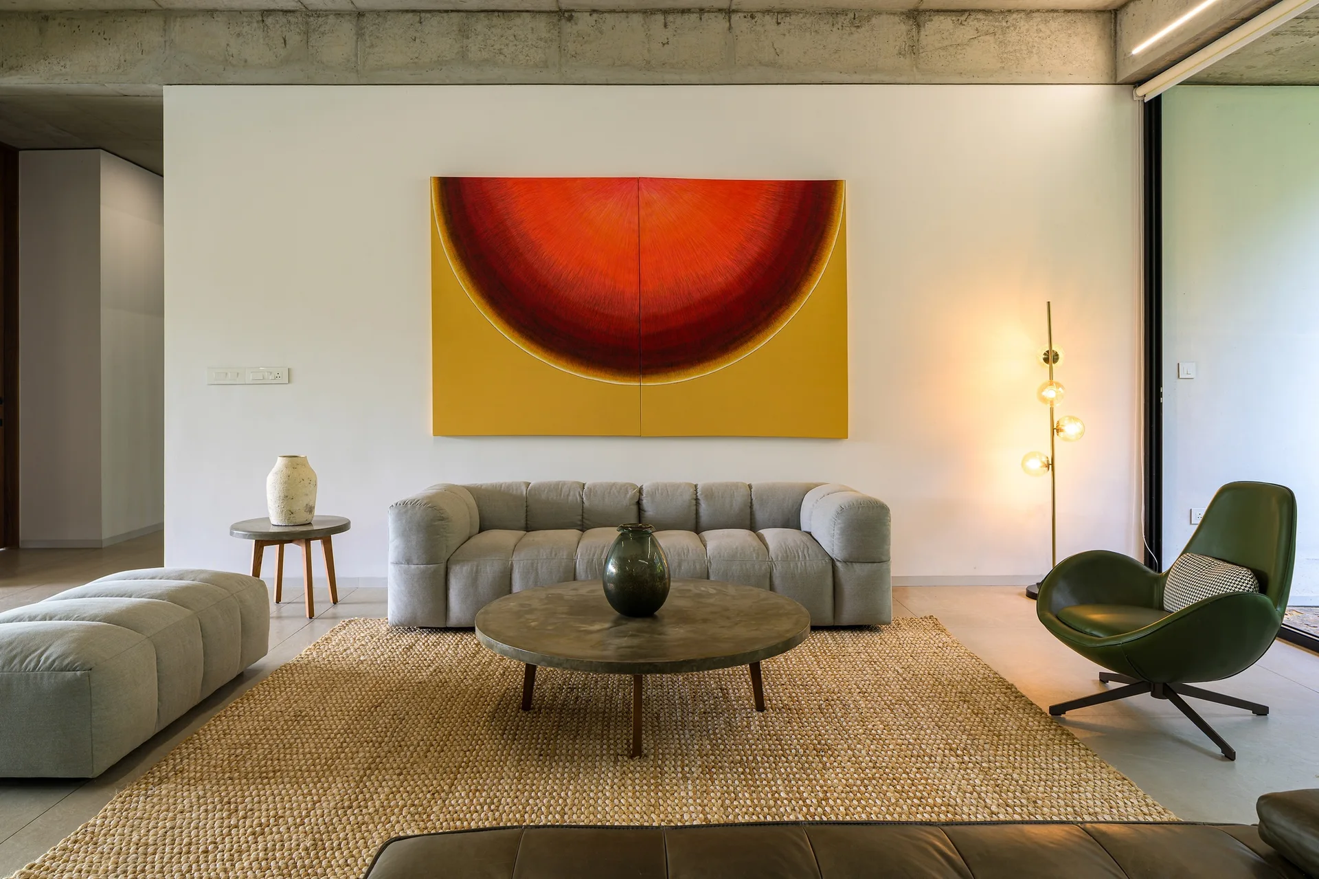 A grey tufted sofa sits beneath a large red and yellow painting in a living room with an exposed concrete ceiling and a textured woven rug.