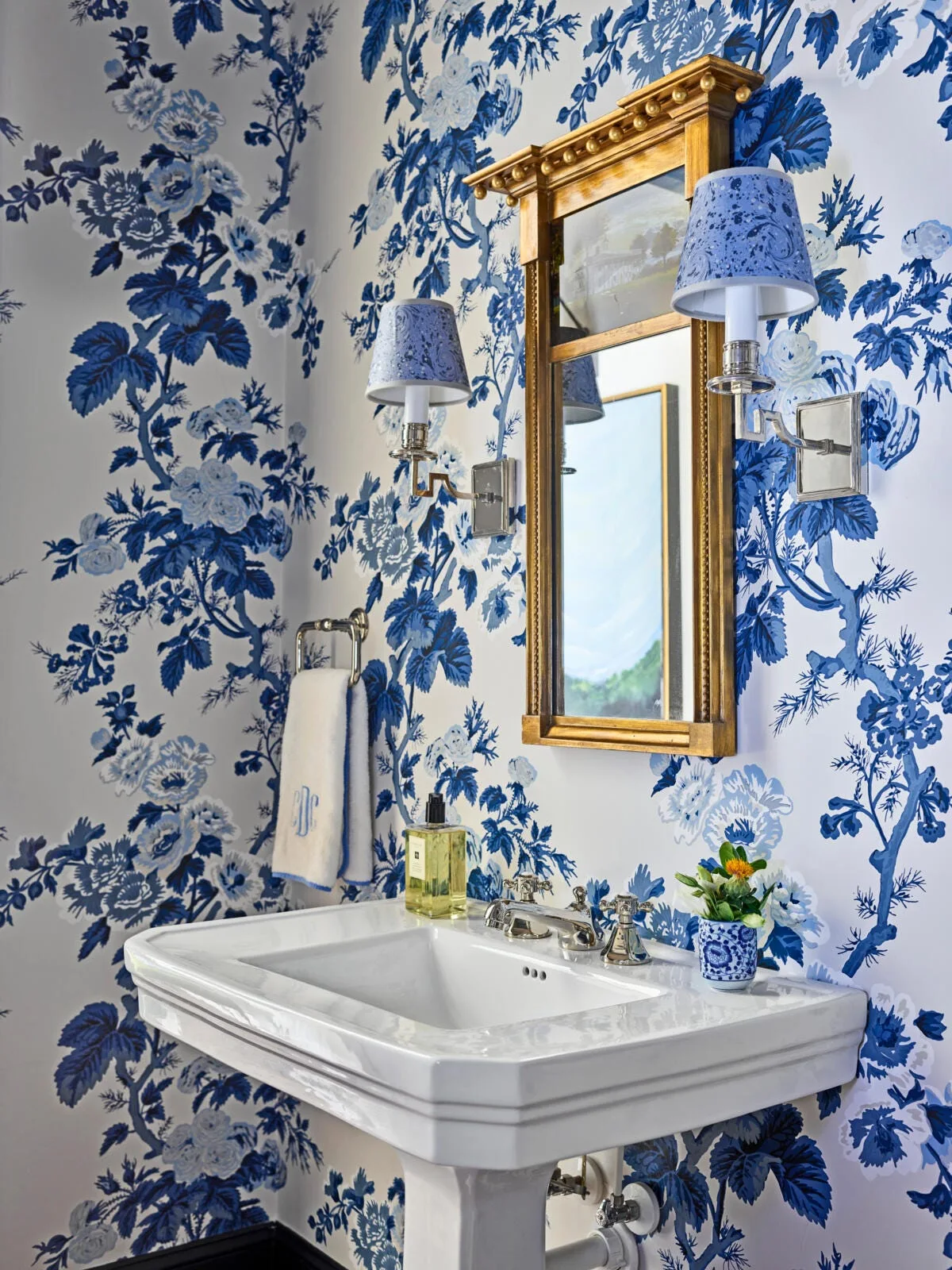 A powder room features bold blue and white floral wallpaper, a white pedestal sink, and a gilded mirror flanked by two wall sconces.