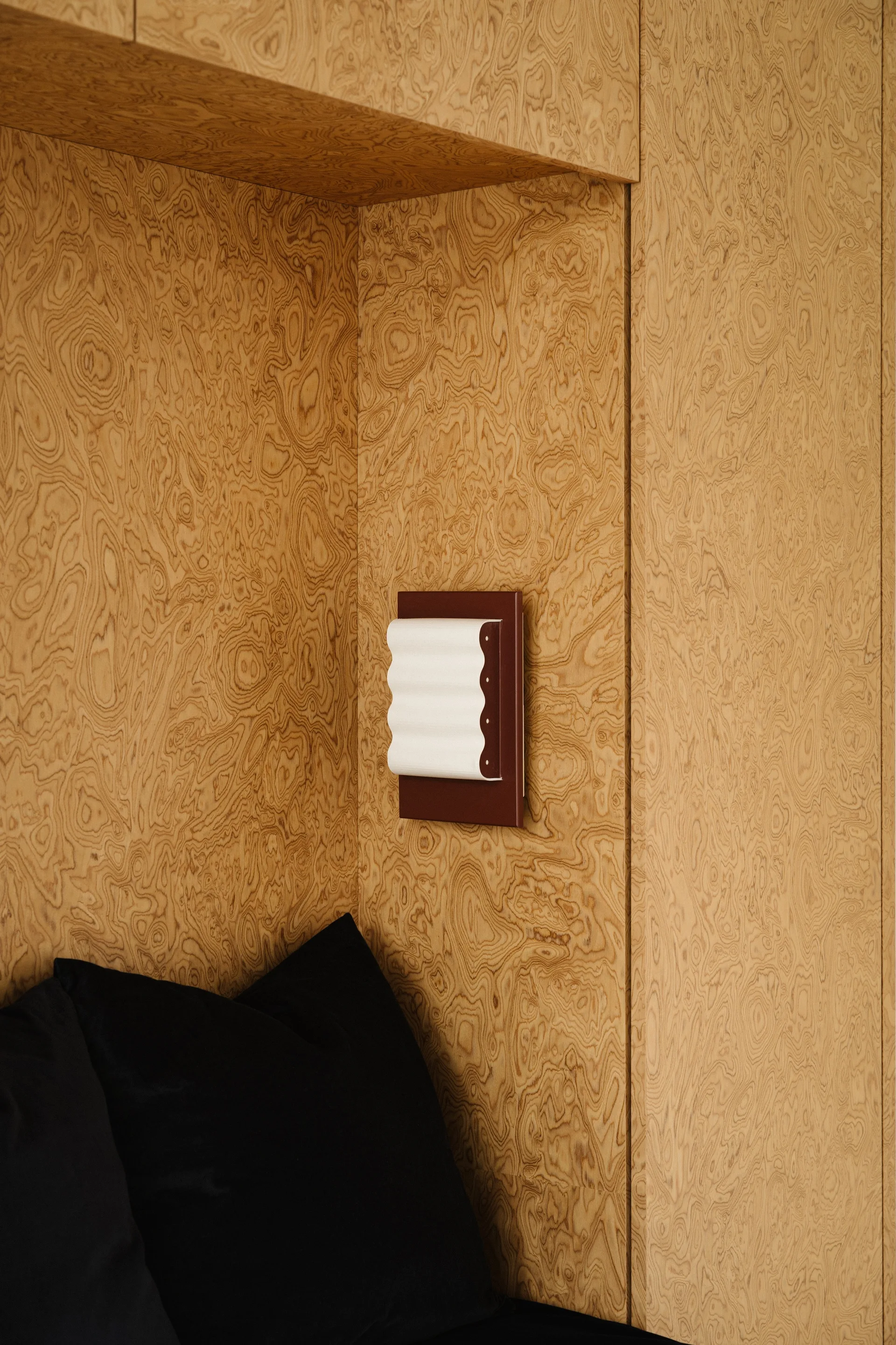 A wavy white wall sconce with a dark red base is mounted on swirling burl wood paneling above black bed pillows.