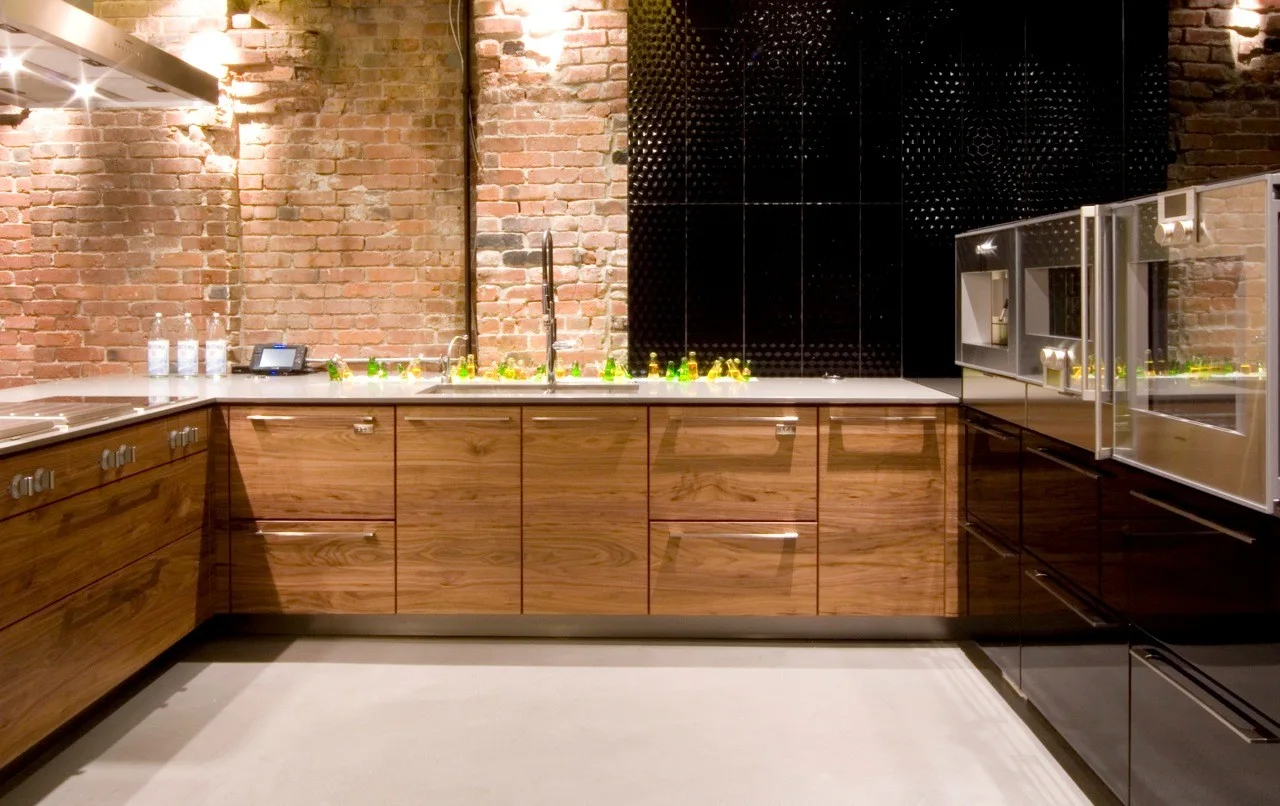 A modern kitchen featuring natural wood cabinets, a light grey floor, an exposed brick wall, and a textured black glossy tile backsplash.