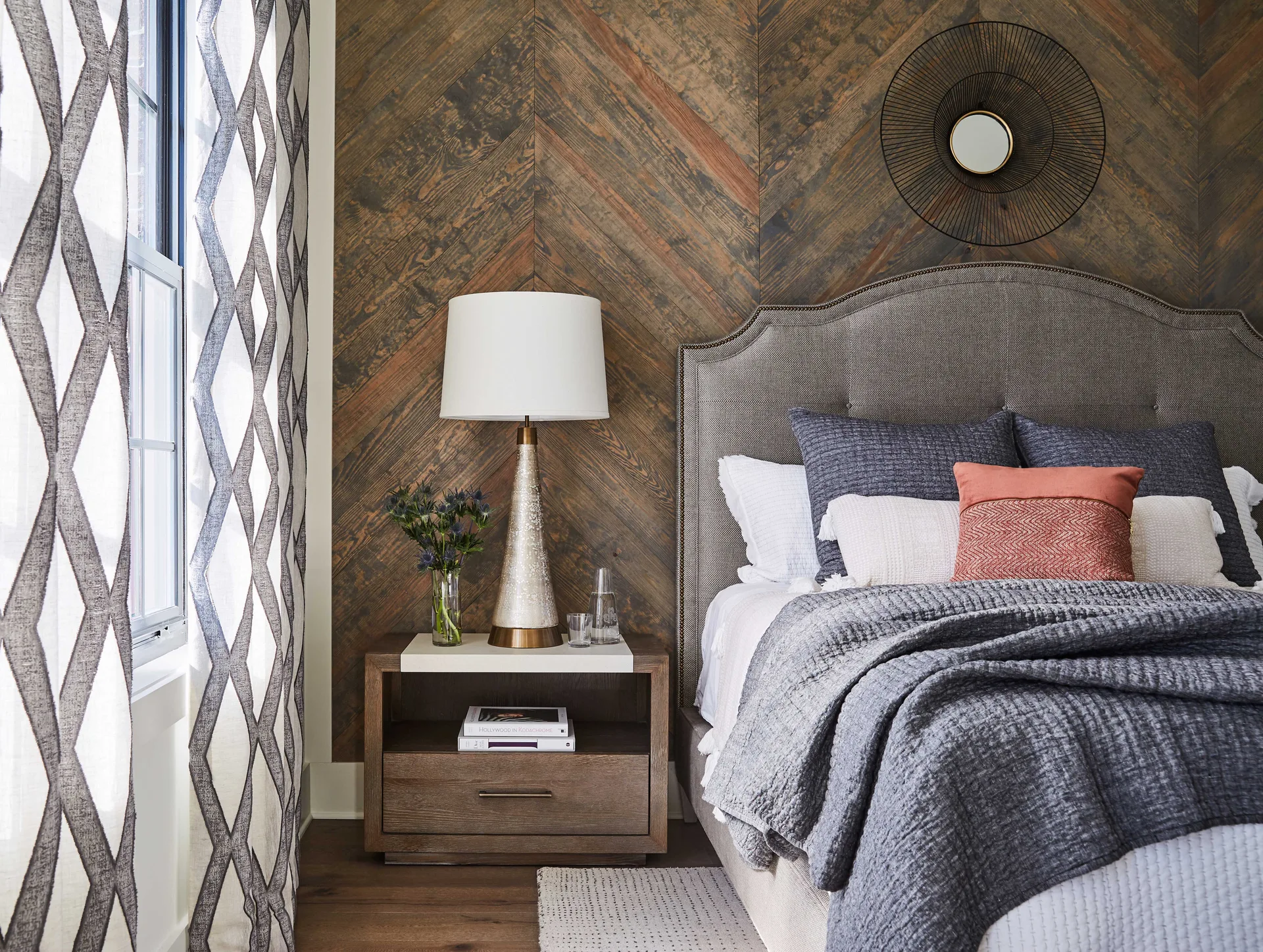 A bedroom displays a grey upholstered headboard against a rustic herringbone wood-paneled wall next to a wooden nightstand and patterned curtain.