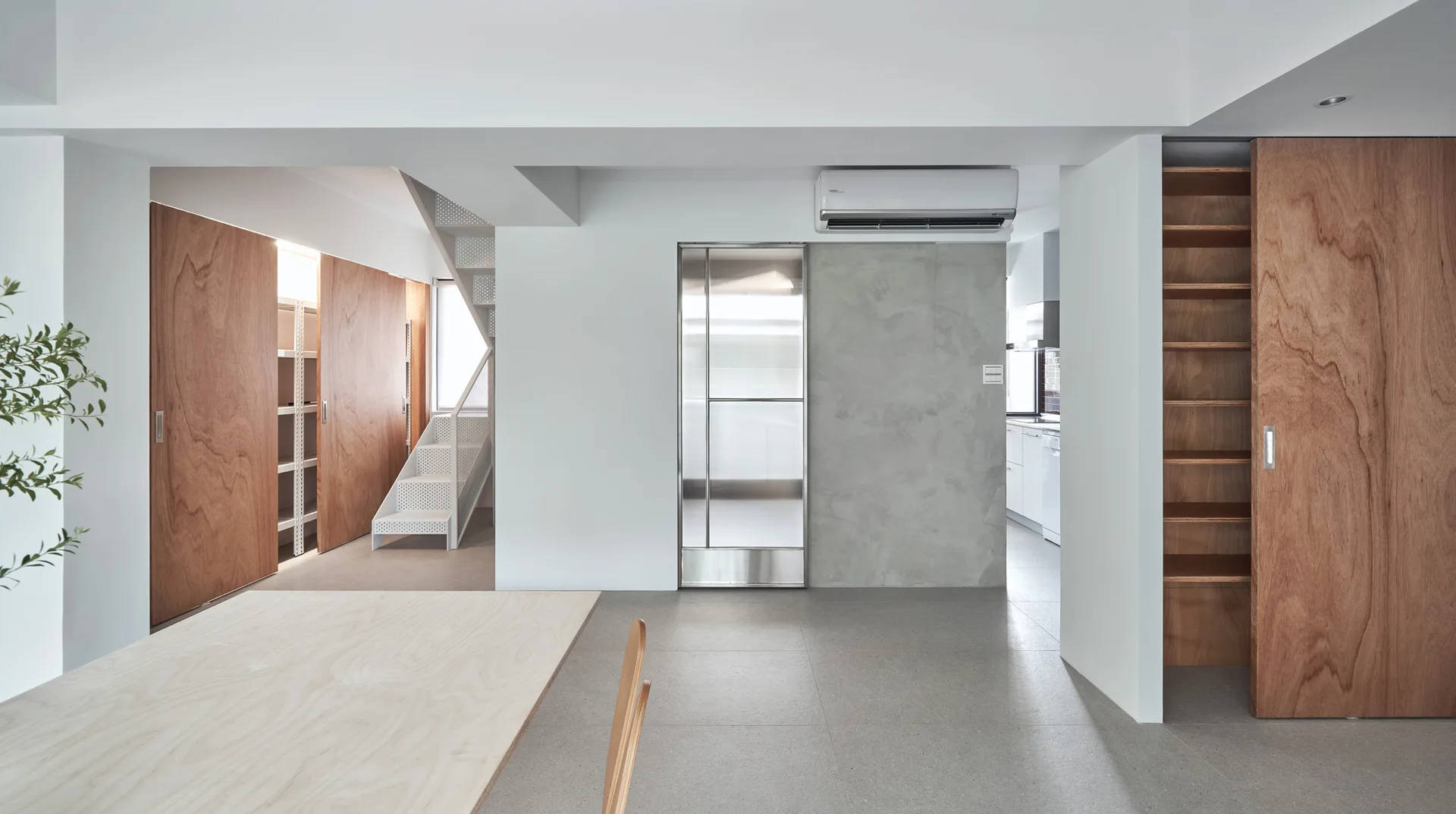 A minimalist open-plan home interior features wood sliding doors, a white perforated metal staircase, gray tiled flooring, and a plywood dining table.