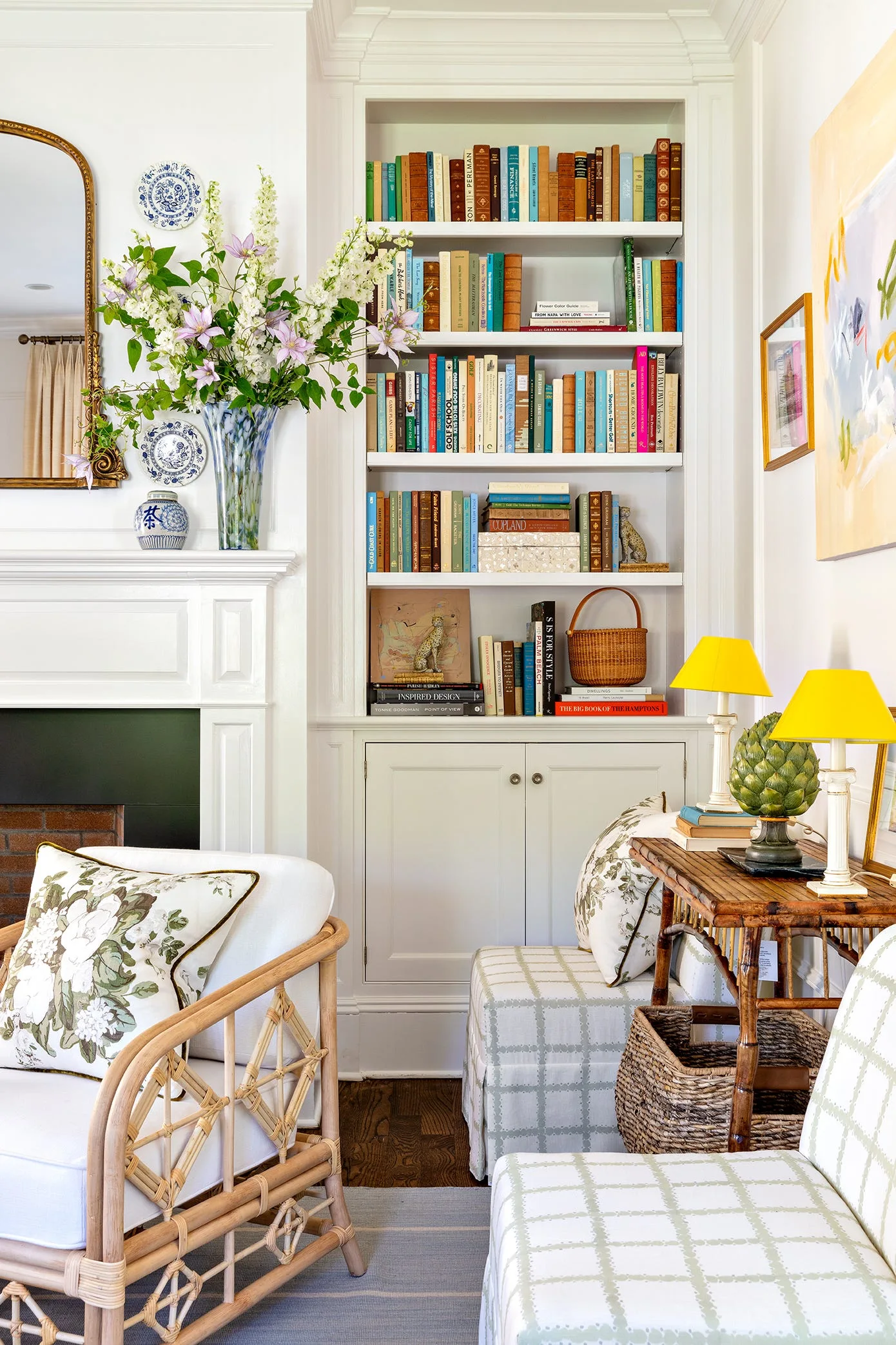 A white built-in bookcase filled with books sits next to a fireplace mantel in a living room with rattan armchairs and a bamboo console table.