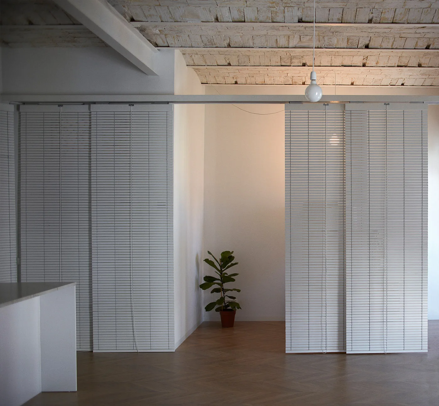 A minimalist interior space with white slatted sliding partition blinds, herringbone wood flooring, and an exposed brick ceiling.