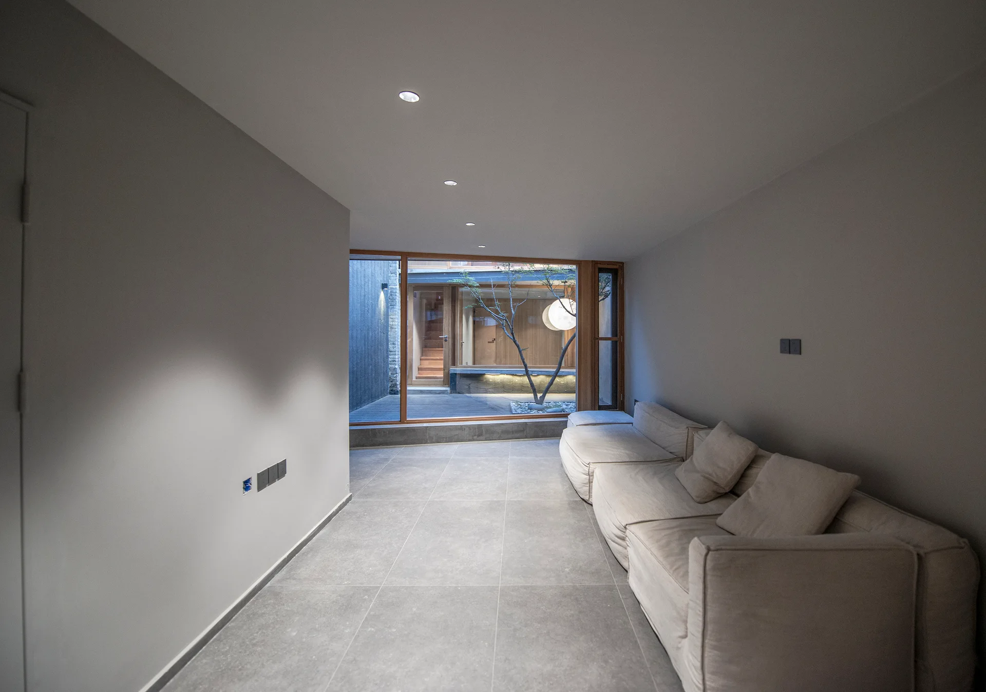 A minimalist grey living room featuring a long beige modular sofa, large-format tile flooring, and a large window looking out onto a central courtyard.