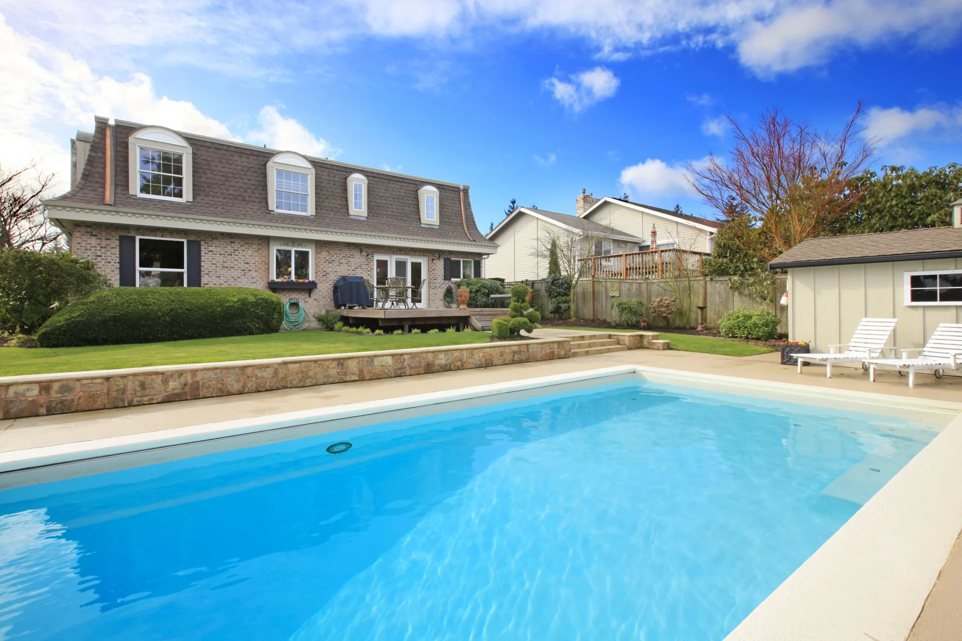 A rectangular swimming pool sits in a residential backyard next to a stone retaining wall, green lawn, and a brick house with a mansard roof.