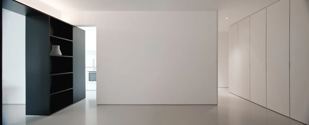 A minimalist white corridor features seamless floor-to-ceiling cabinetry on one side and a matte black open-shelving unit on the other.