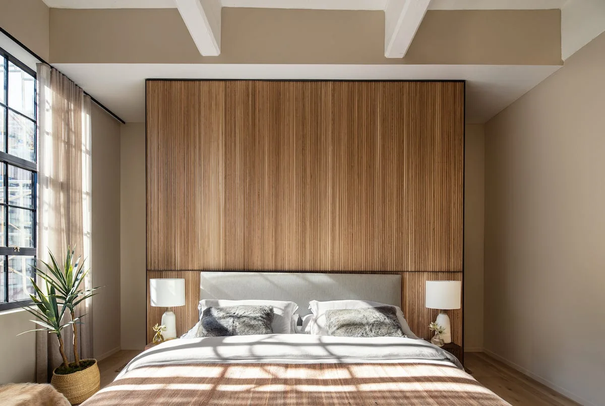A modern bedroom features a large vertical wood slat accent wall behind a bed flanked by white table lamps and a window with sheer curtains.