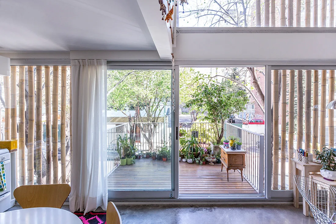 Wide sliding glass doors open to a wood-decked balcony filled with plants, framed by a vertical bamboo privacy screen and sheer white curtains.
