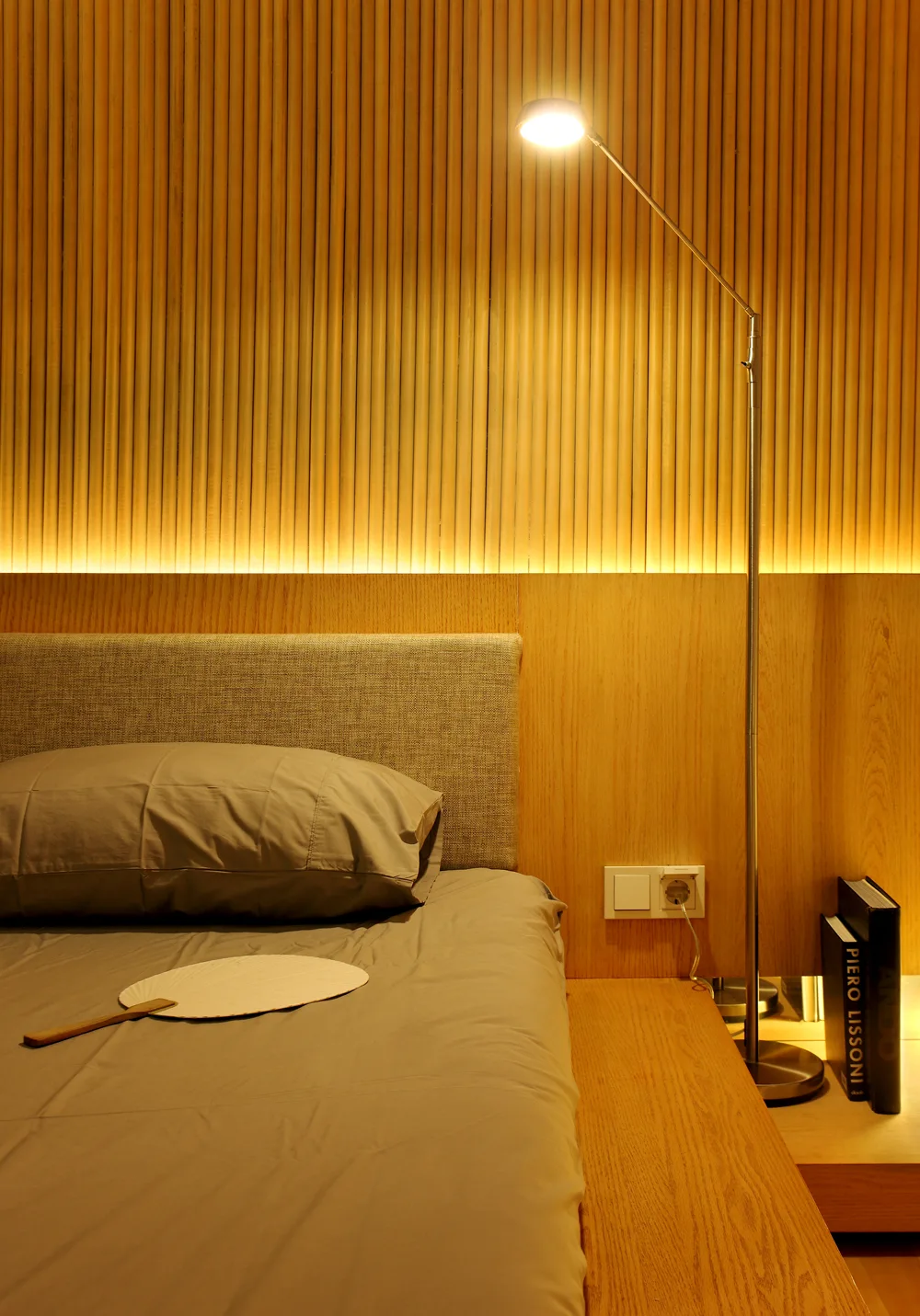 A bed with grey linens and an upholstered headboard sits against a warm wood slatted wall illuminated by an integrated LED strip and a tall metal floor lamp.