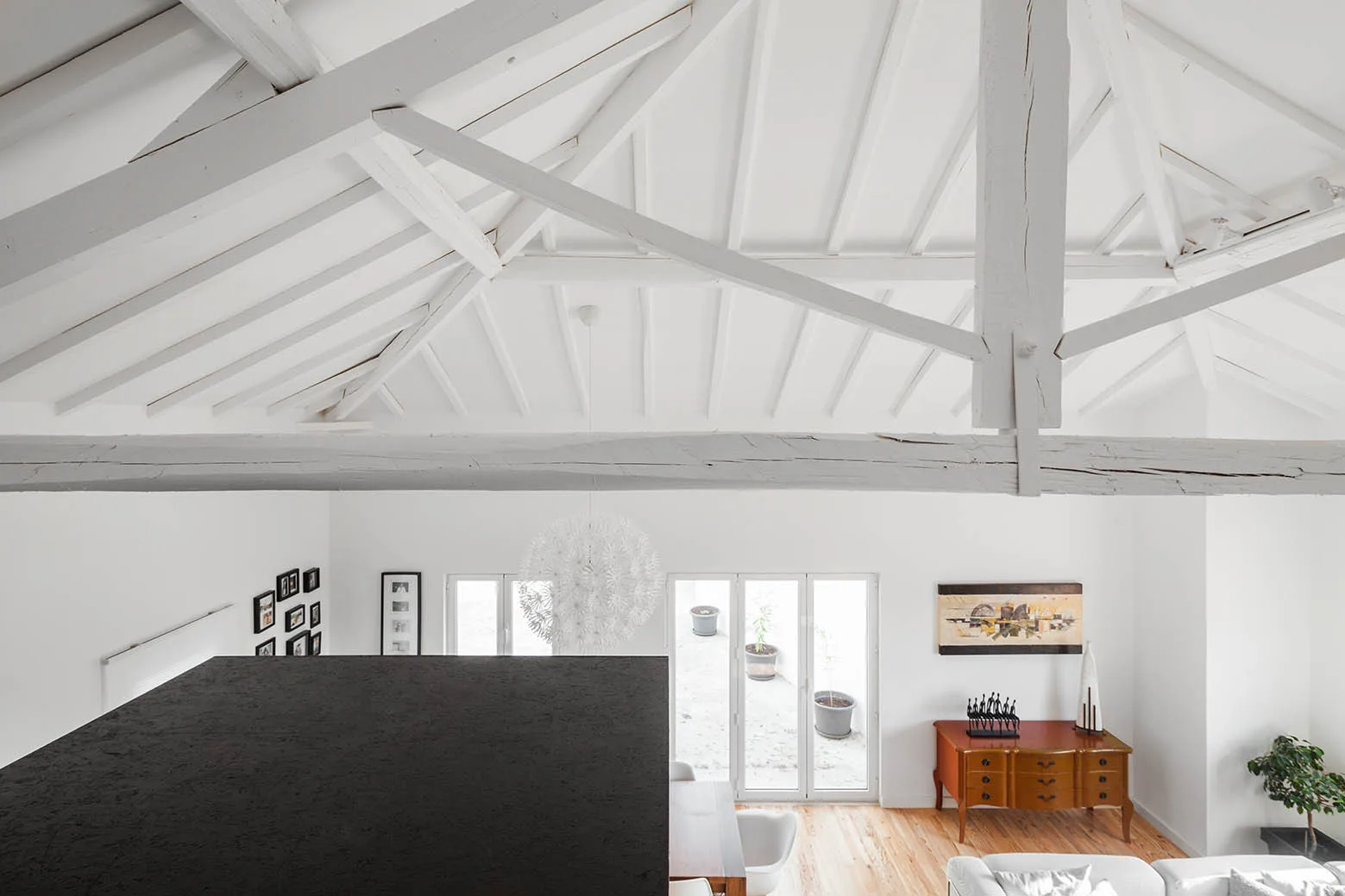 A high-angle view looking down from a loft onto a white living room with high vaulted ceilings, white painted wooden beams, and a wooden sideboard.