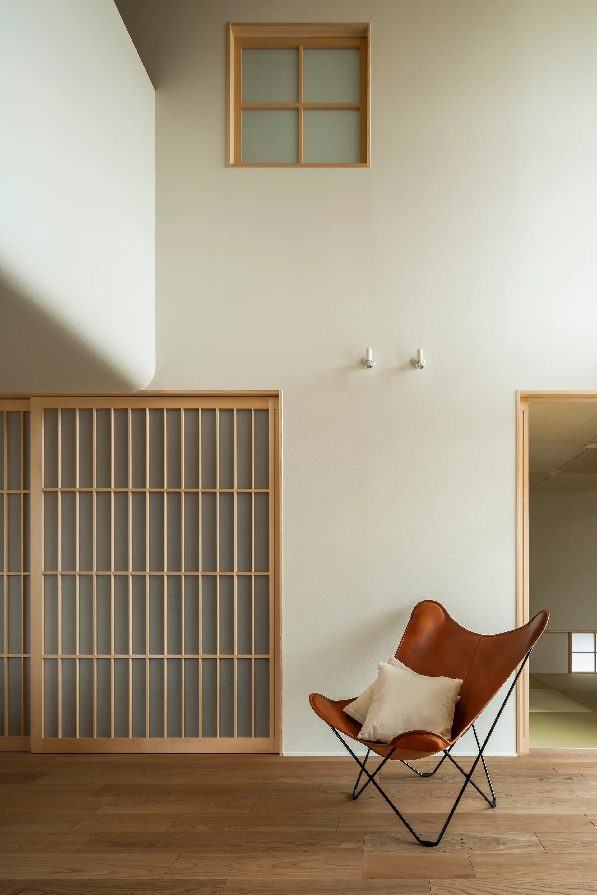 A brown leather butterfly chair sits on a hardwood floor in a minimalist room with shoji sliding doors and high off-white walls.