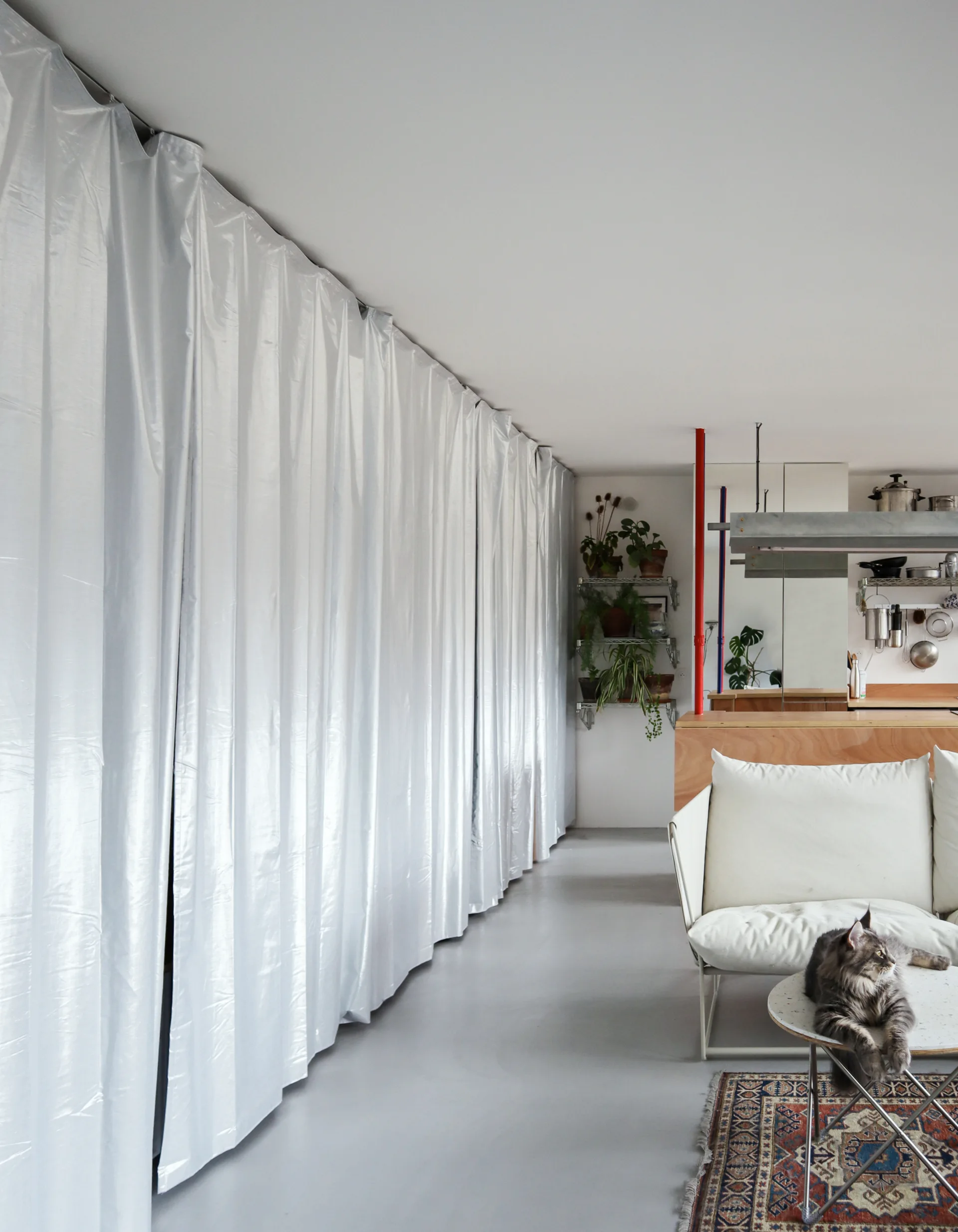 A modern living room features a full-length reflective white curtain wall, a white sofa, a smooth grey concrete floor, and kitchen cabinetry in the background.