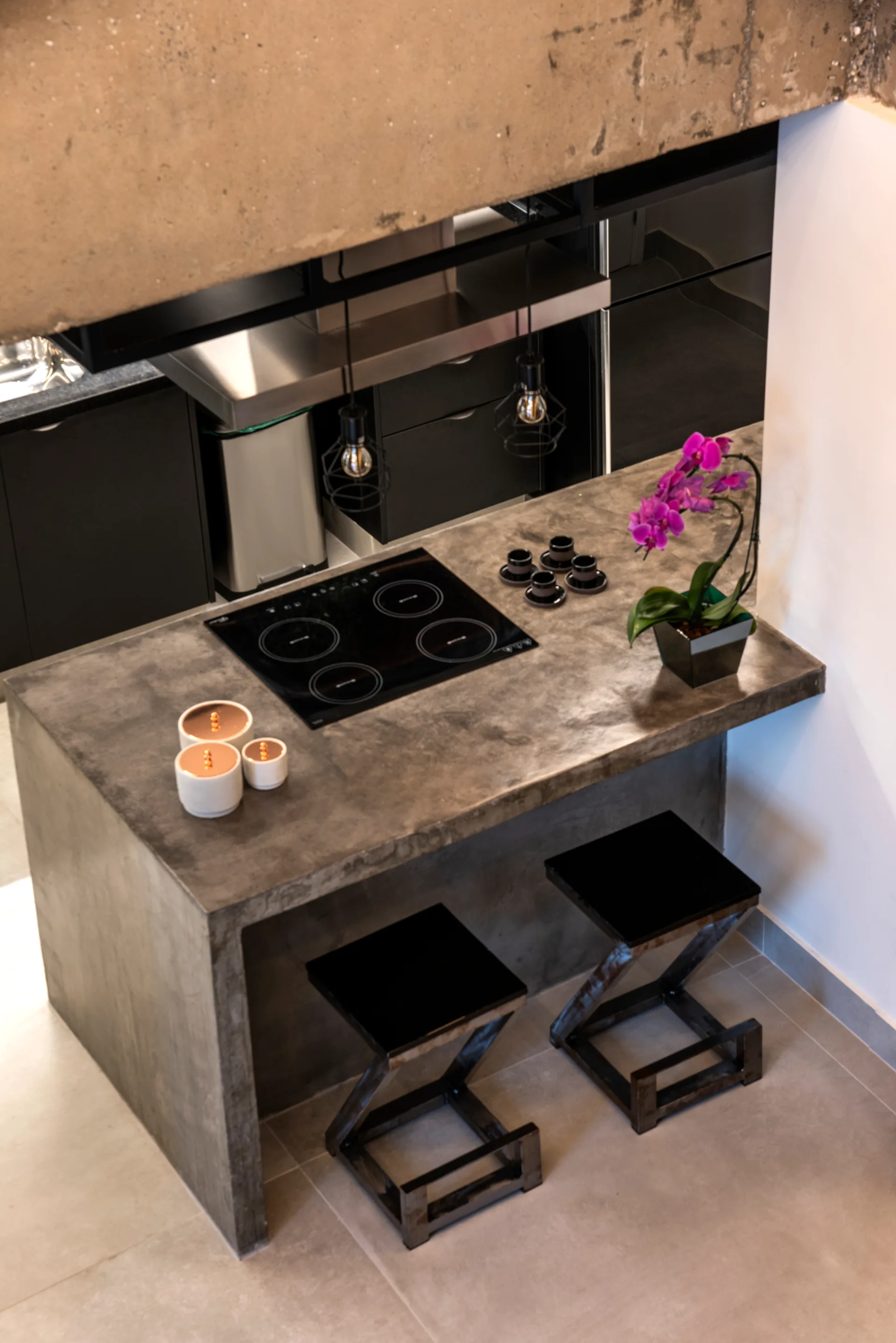 A high-angle view of a grey concrete kitchen island featuring a black cooktop, flanked by two black zig-zag metal bar stools and dark cabinetry.