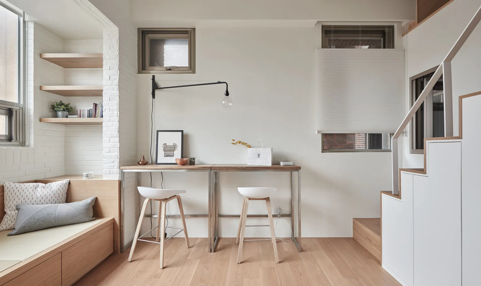 A light-filled room features a long wooden console desk with two stools, a built-in bench under floating shelves, and stairs with white storage drawers.