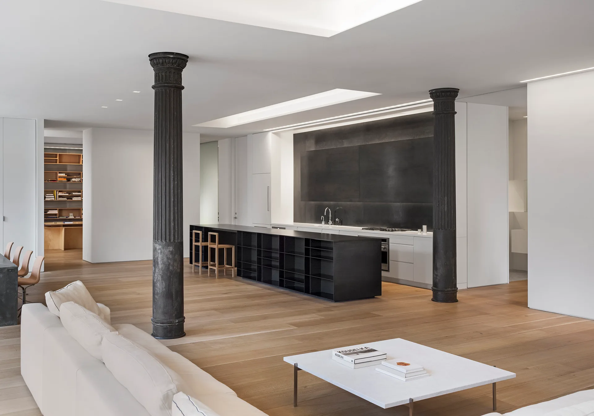A spacious open loft features a minimalist kitchen with a black island and cabinetry flanked by two black fluted columns over light hardwood floors.