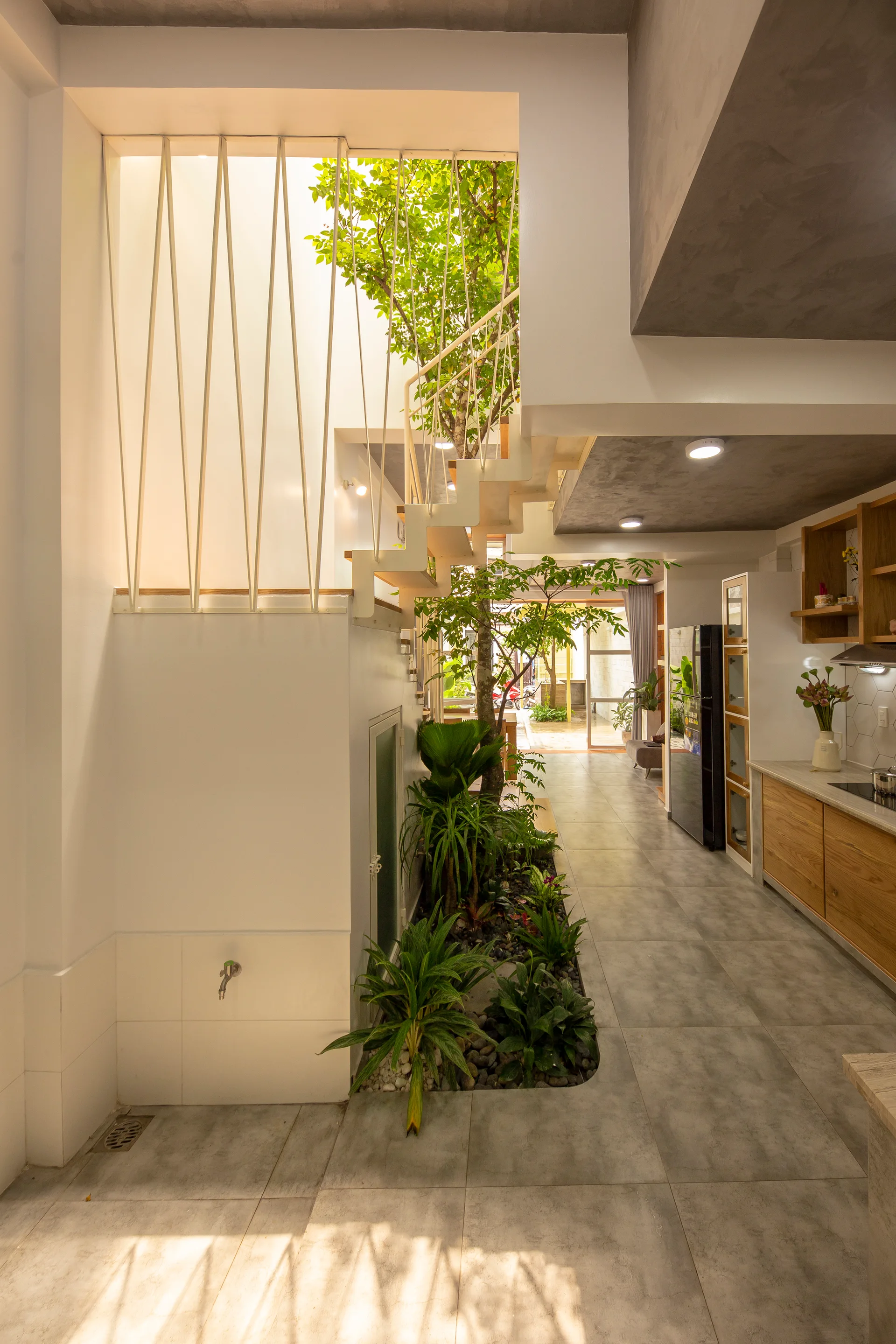 A modern home interior features an indoor garden planted beneath a white staircase, adjacent to a kitchen with grey tile floors.