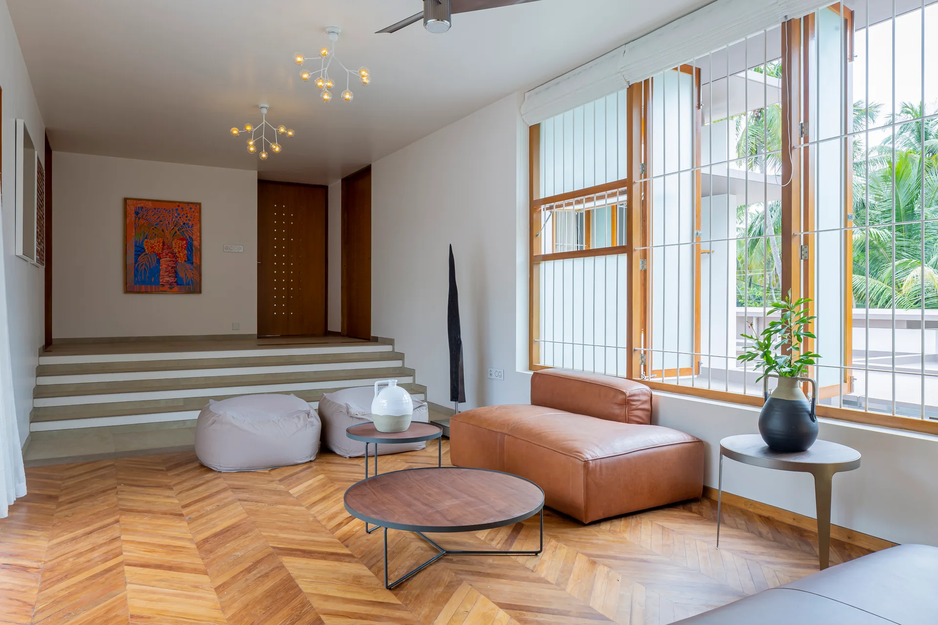 A modern living room features a brown modular leather chair, round nesting coffee tables, a herringbone wood floor, and steps leading to a raised platform.