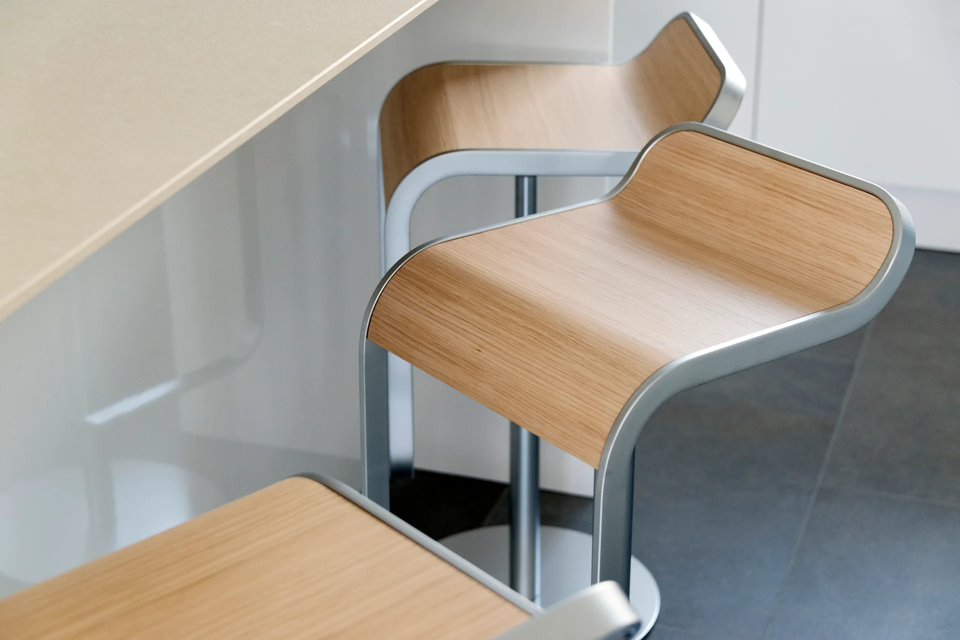 Two modern bar stools featuring curved light wood seats and silver metal frames stand next to a white kitchen counter on a dark gray tile floor.