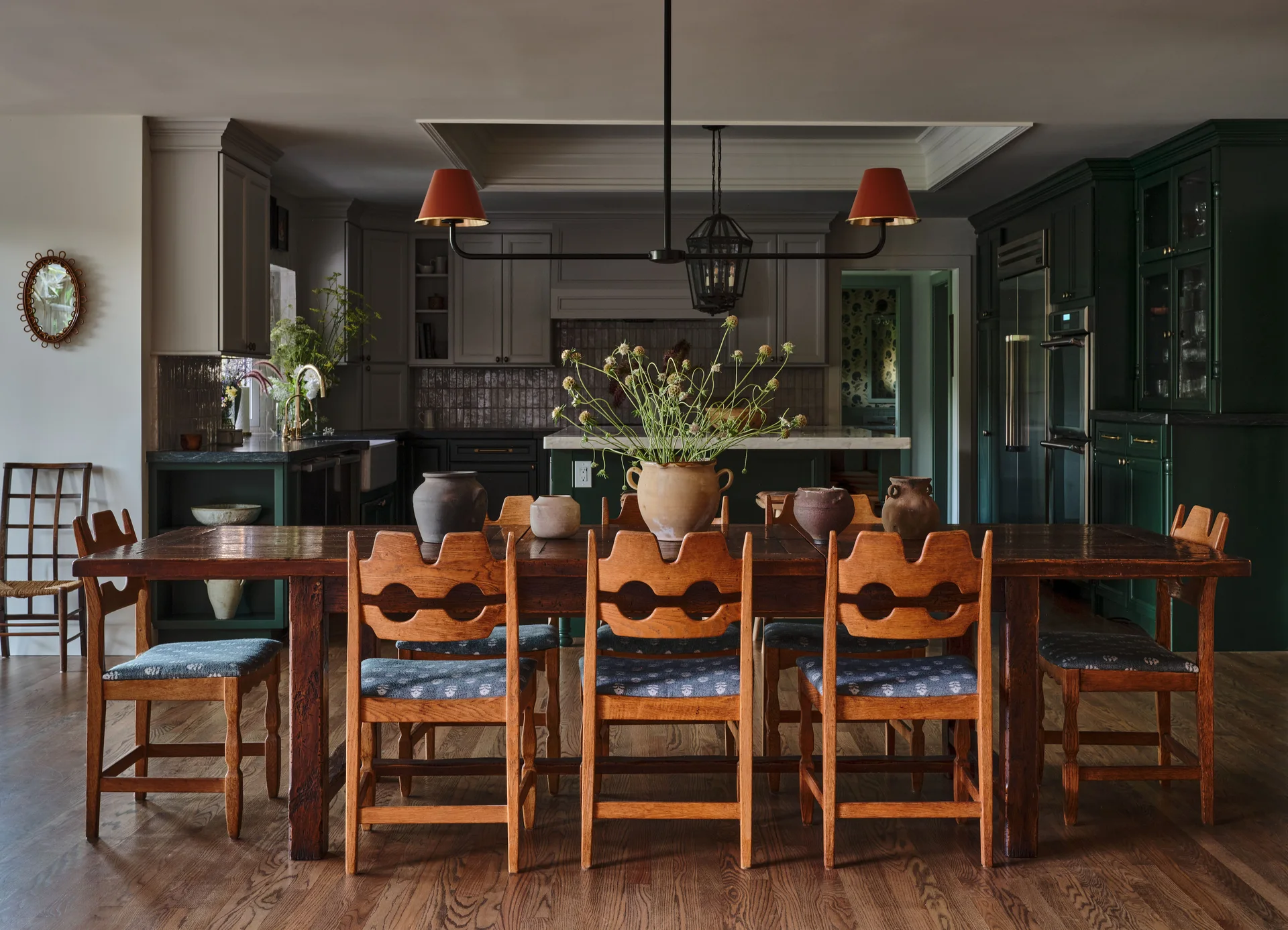 A rustic wooden dining table with unique cutout chairs is set in a home dining space with dark green kitchen cabinets in the background.