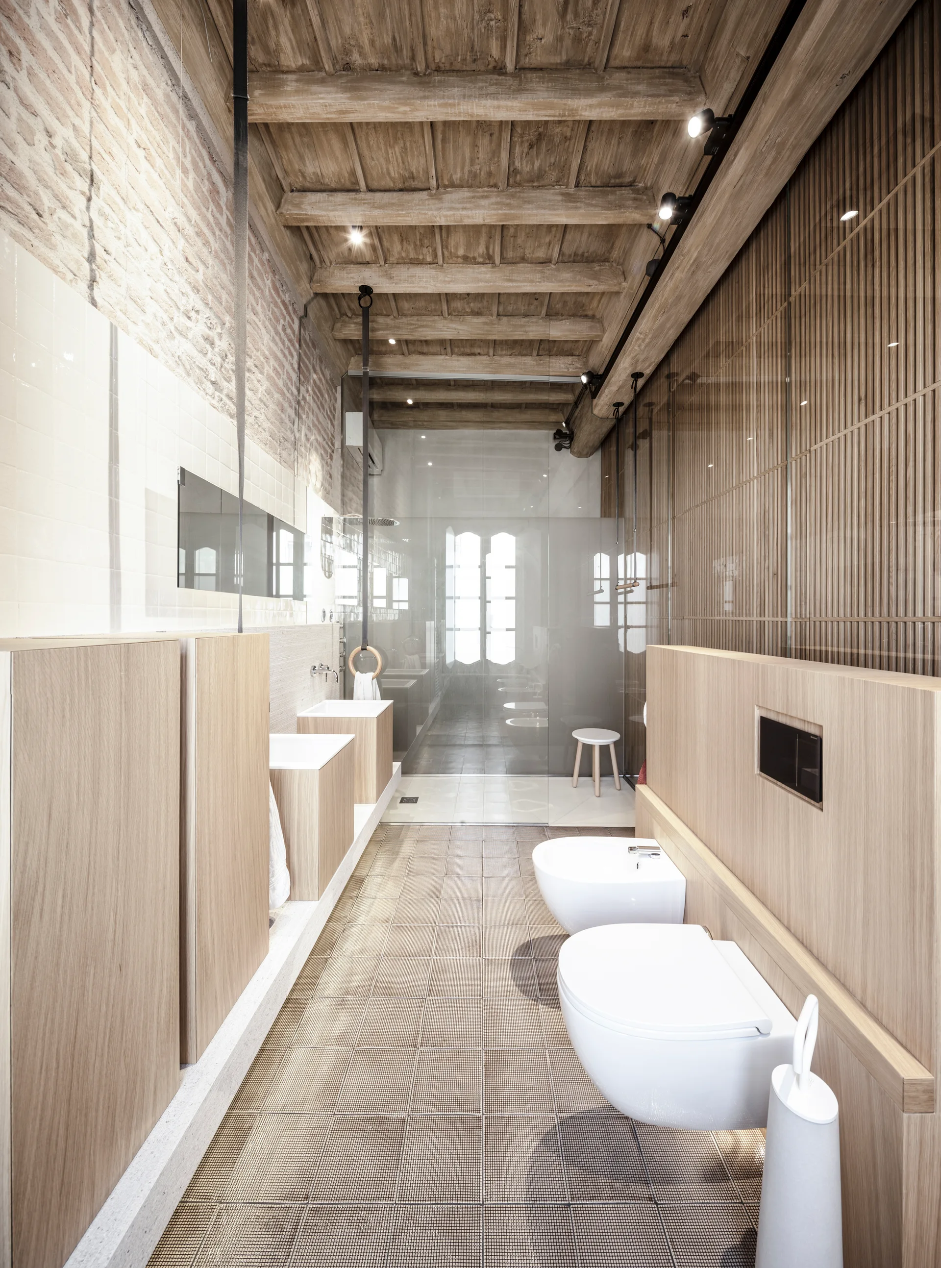 A narrow bathroom featuring wall-mounted ceramic fixtures, light wood vanities, an exposed brick wall, and rustic wood ceiling beams.