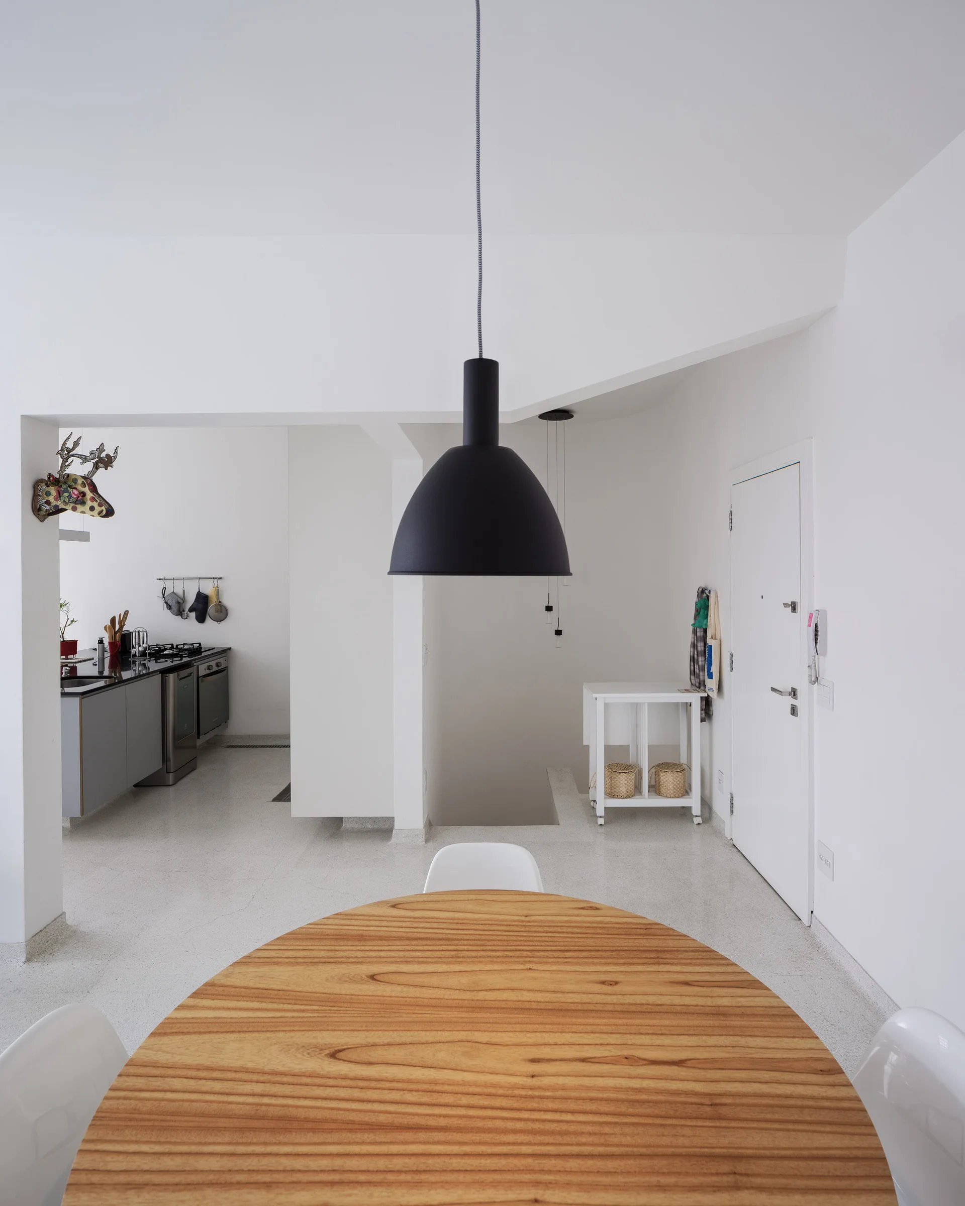 A minimalist dining space features a circular wooden table under a large black dome pendant light, with an open white kitchen visible in the background.