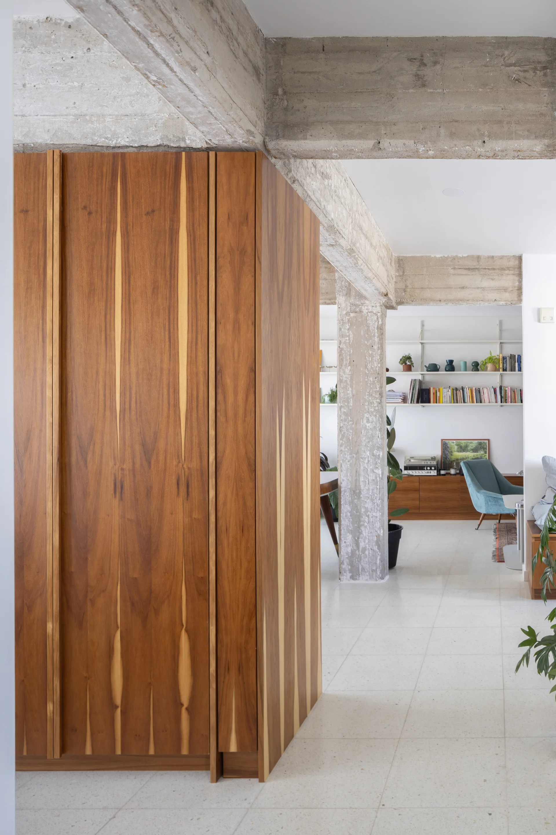 An interior view showing a large warm wood cabinet next to exposed concrete columns, with a bright living area and shelving in the background.