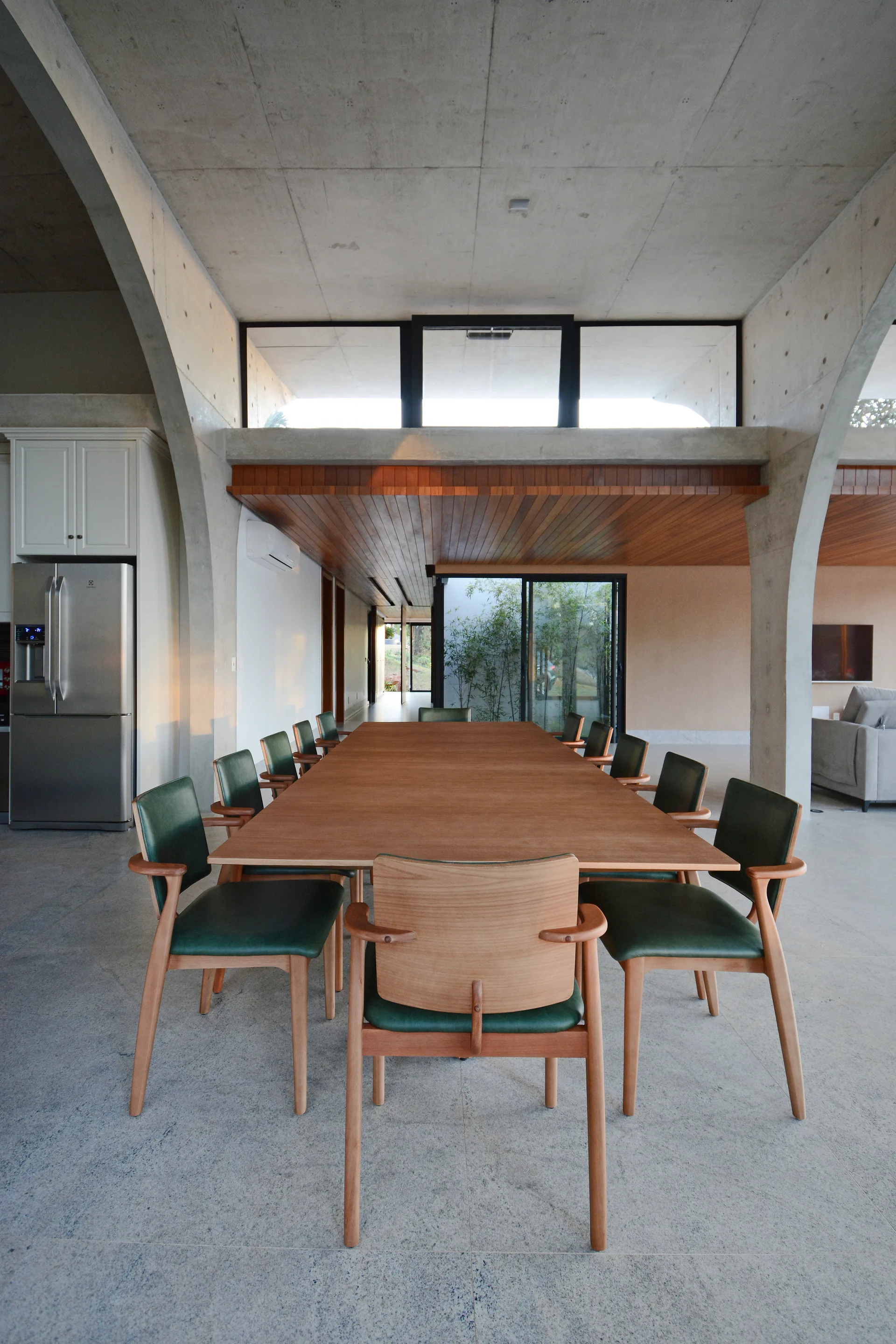 A long wooden dining table and green leather chairs stand under soaring raw concrete arches in a modern open-plan home.