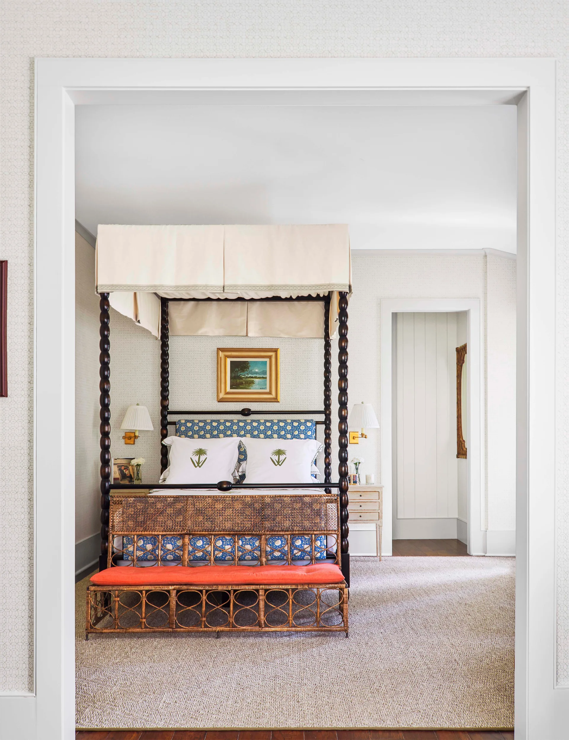 A bedroom viewed through a doorway features a dark wood spindle four-poster bed with blue patterned bedding and a wicker bench with an orange cushion.