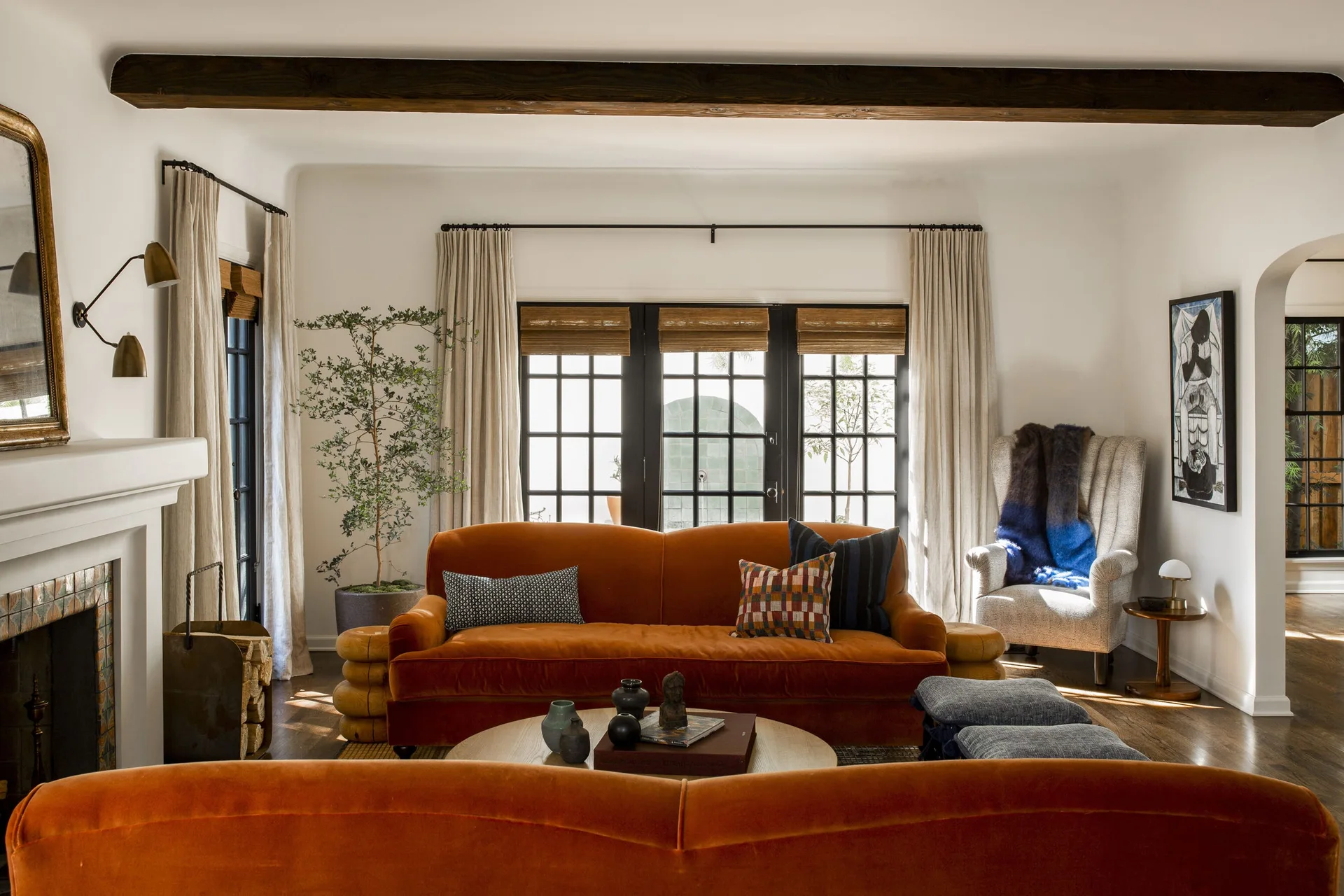 A warm living room features orange velvet sofas, a dark wood ceiling beam, and a tiled fireplace beneath off-white walls.