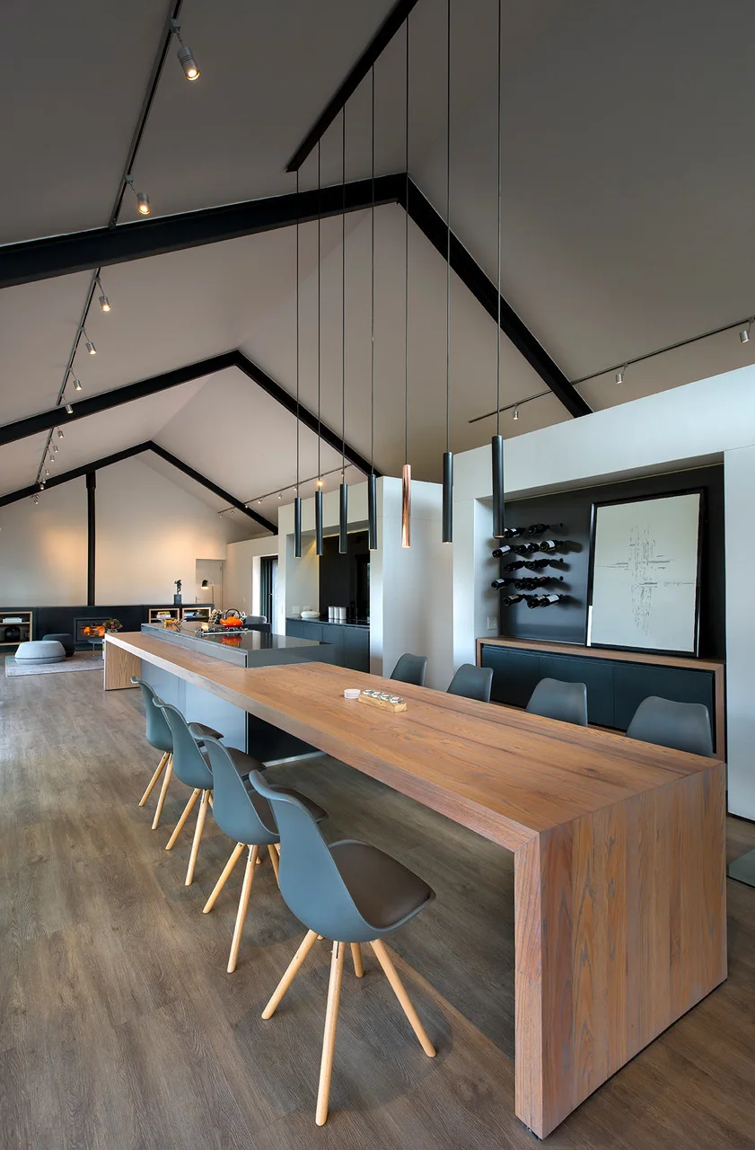 A modern kitchen features a long wooden dining table integrated with the island under a vaulted ceiling with black beams and hanging pendant lights.