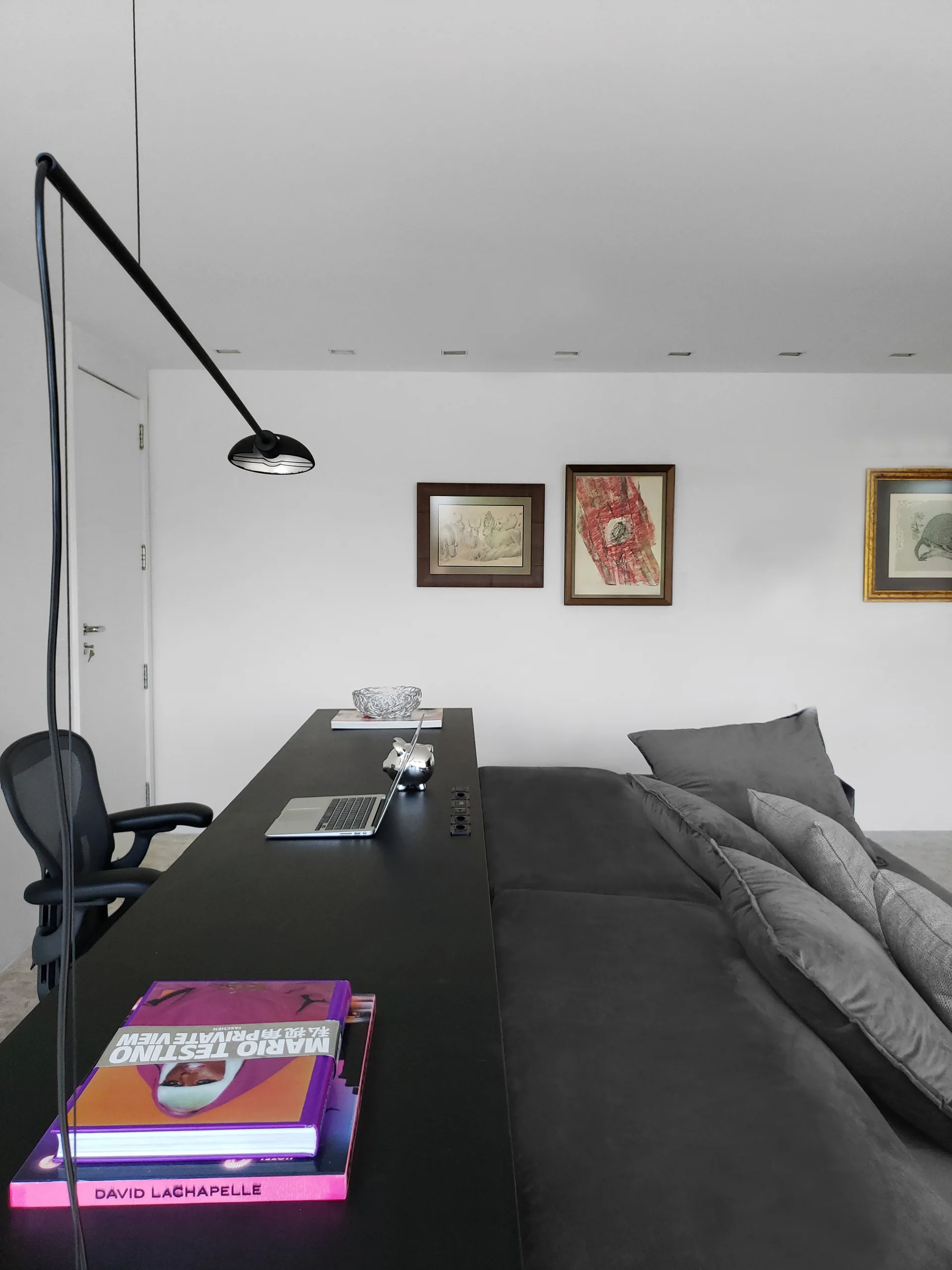 A long black desk sits behind a gray upholstered sofa in a minimalist room with white walls, framed art, and a black arched lamp.