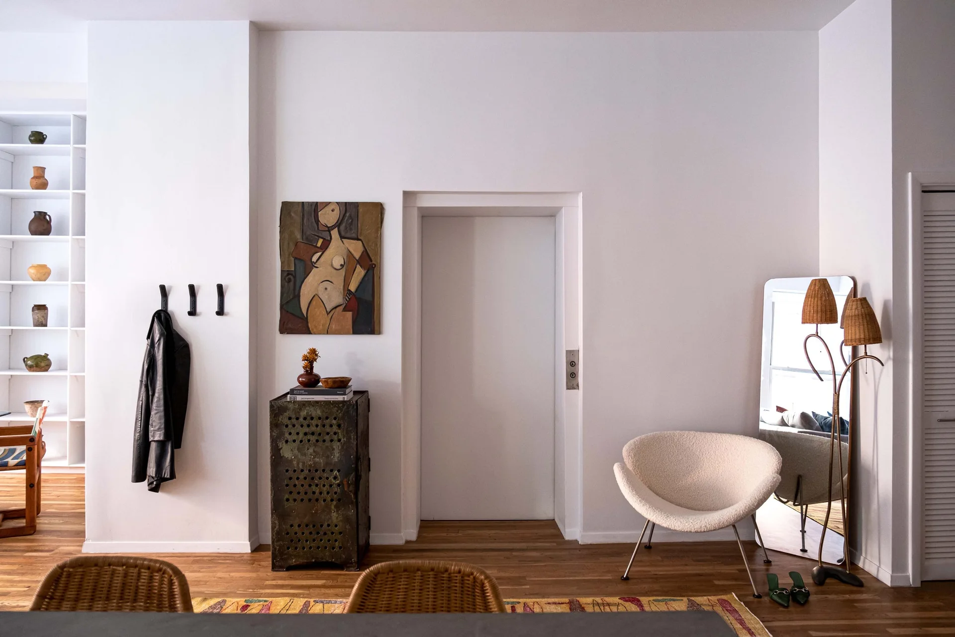 A white entryway features an industrial metal cabinet, a curved bouclé accent chair, and a leaning floor mirror on hardwood floors.