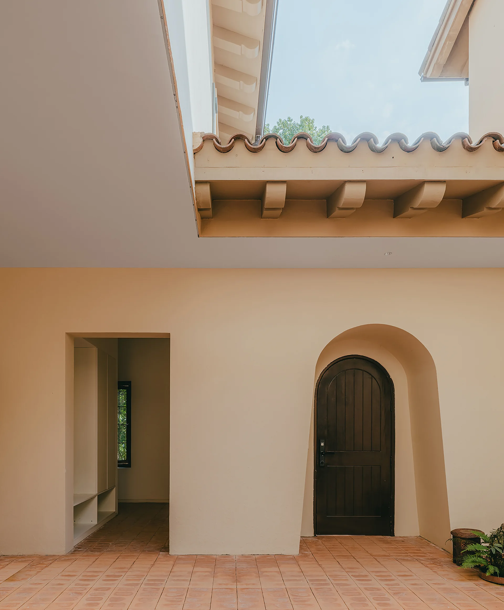 A covered Spanish-style courtyard features a dark arched wood door, beige stucco walls, exposed roof beams, and terracotta tile flooring.