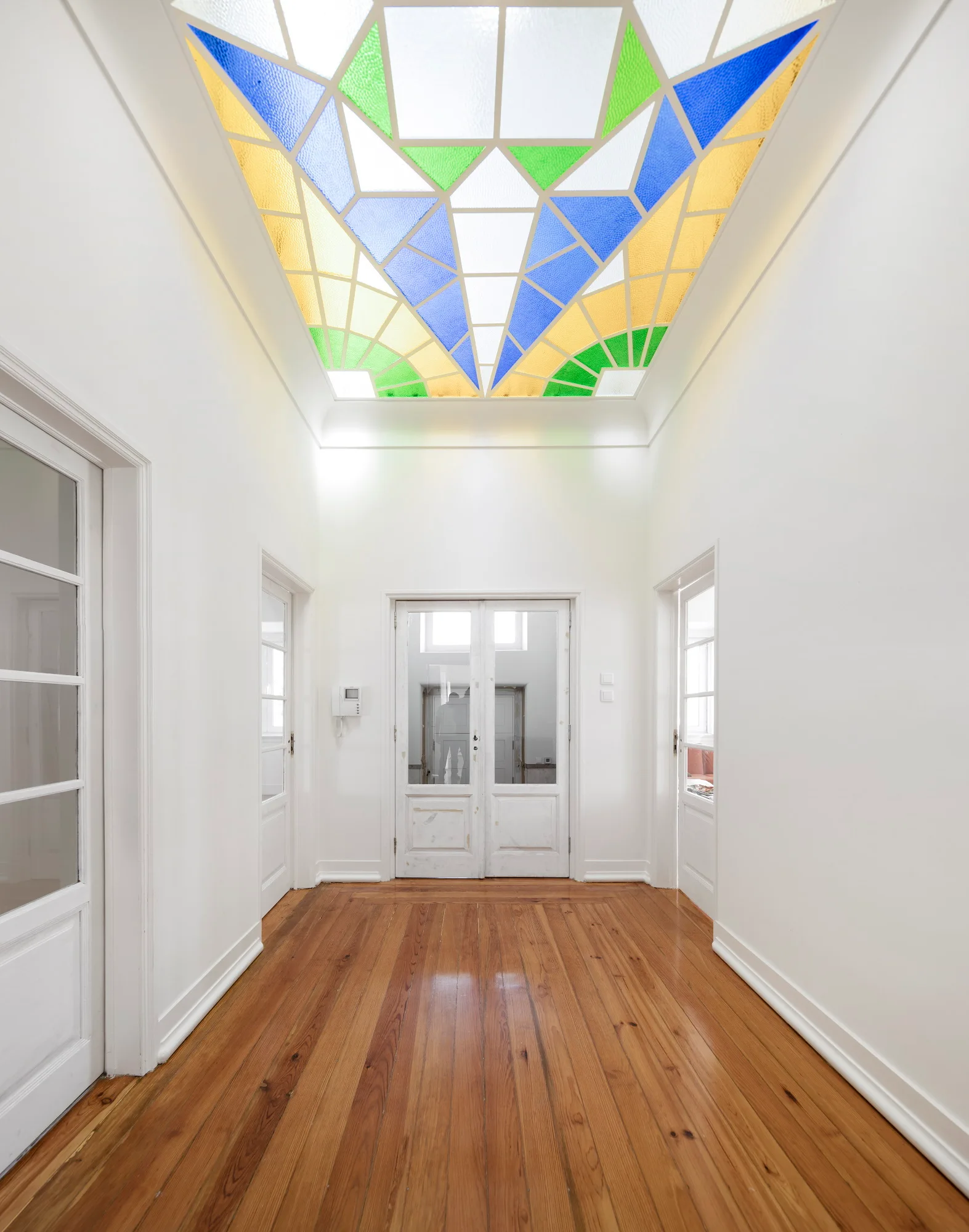 A bright white hallway with polished wood flooring, glass-paneled white doors, and a colorful geometric stained glass ceiling light.