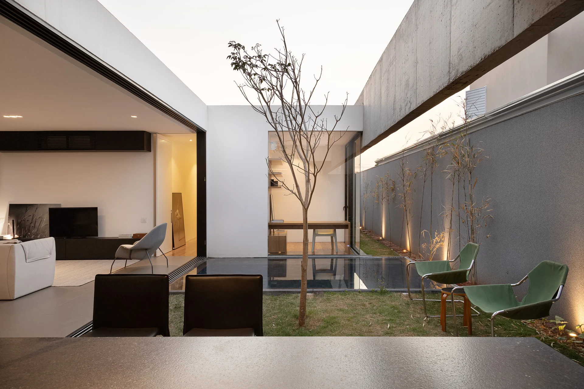 A modern minimalist courtyard features a small reflecting pool, green sling chairs, and open sliding glass doors leading to interior living spaces.