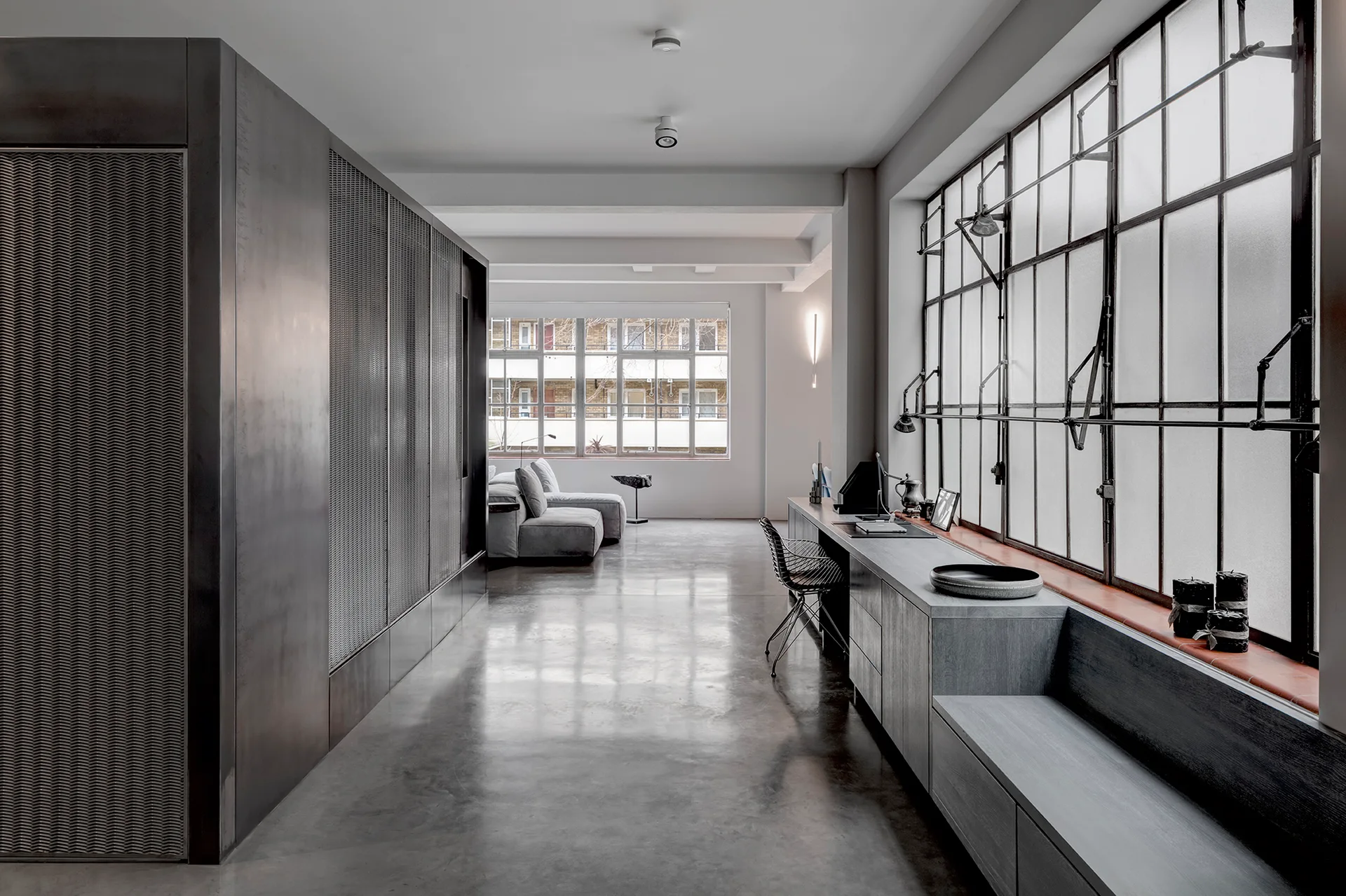 A modern loft workspace features a long built-in desk below large steel-framed windows next to a metal mesh partition and polished concrete floor.