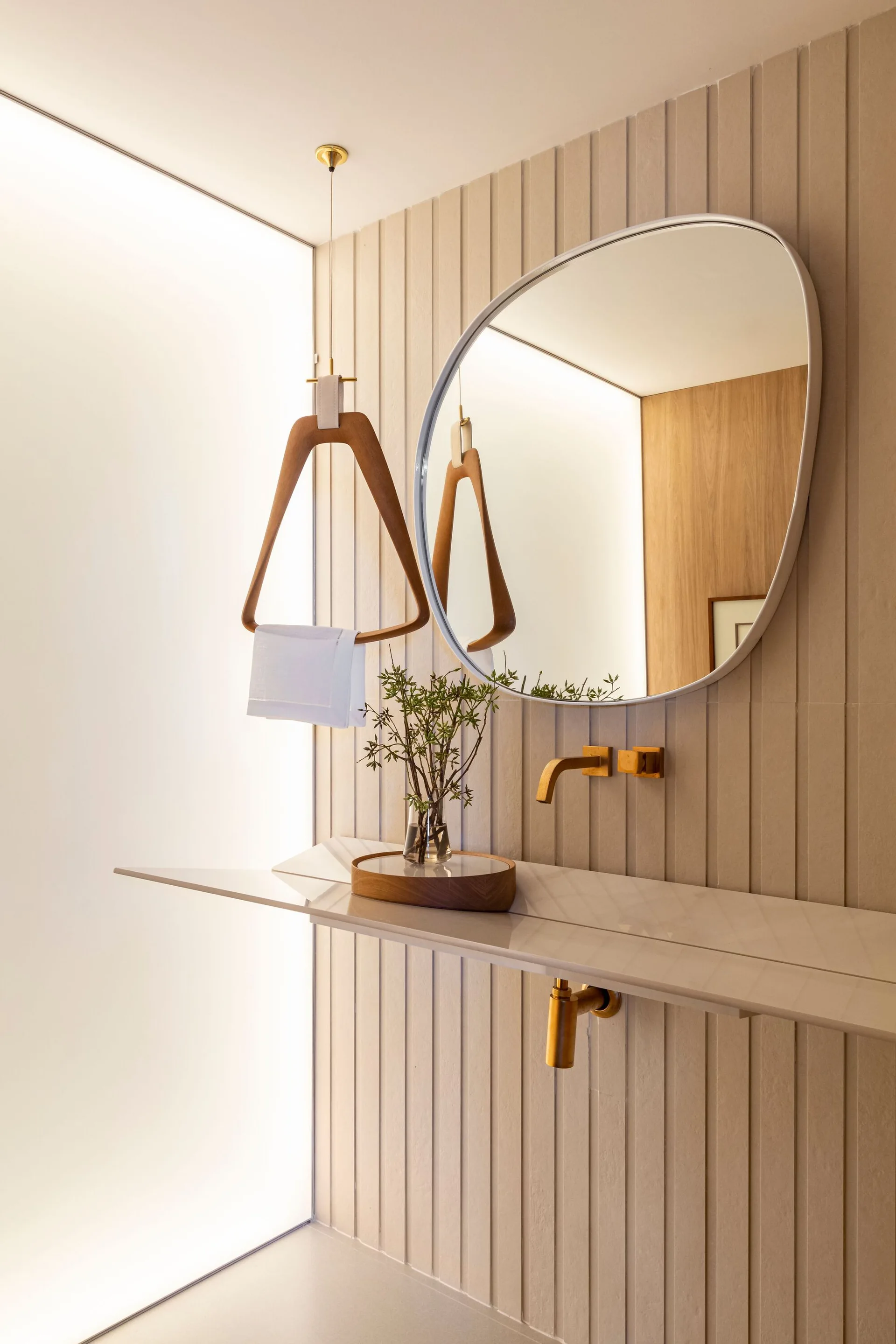 A minimalist beige powder room features an organic-shaped mirror, a wall-mounted gold faucet, and a wooden towel hanger suspended against a grooved wall.