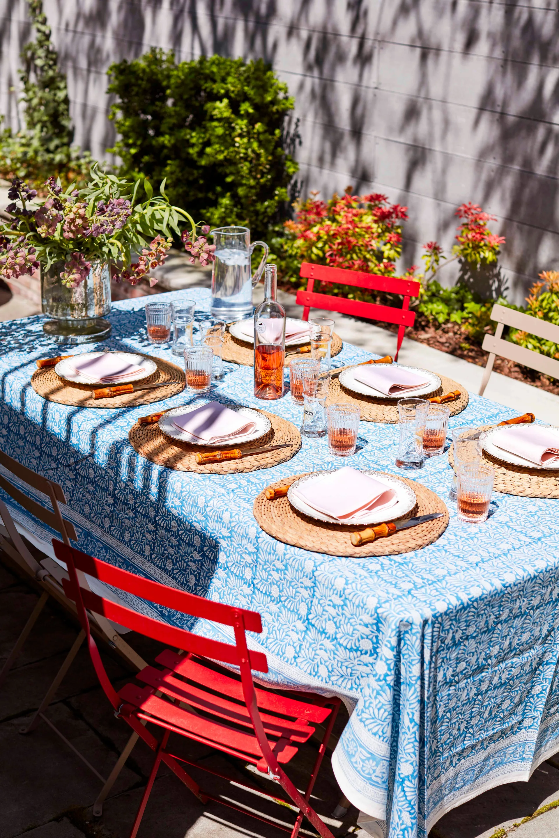 An outdoor dining table is set with a blue floral tablecloth, woven placemats, pink napkins, and red metal folding chairs on a stone patio.