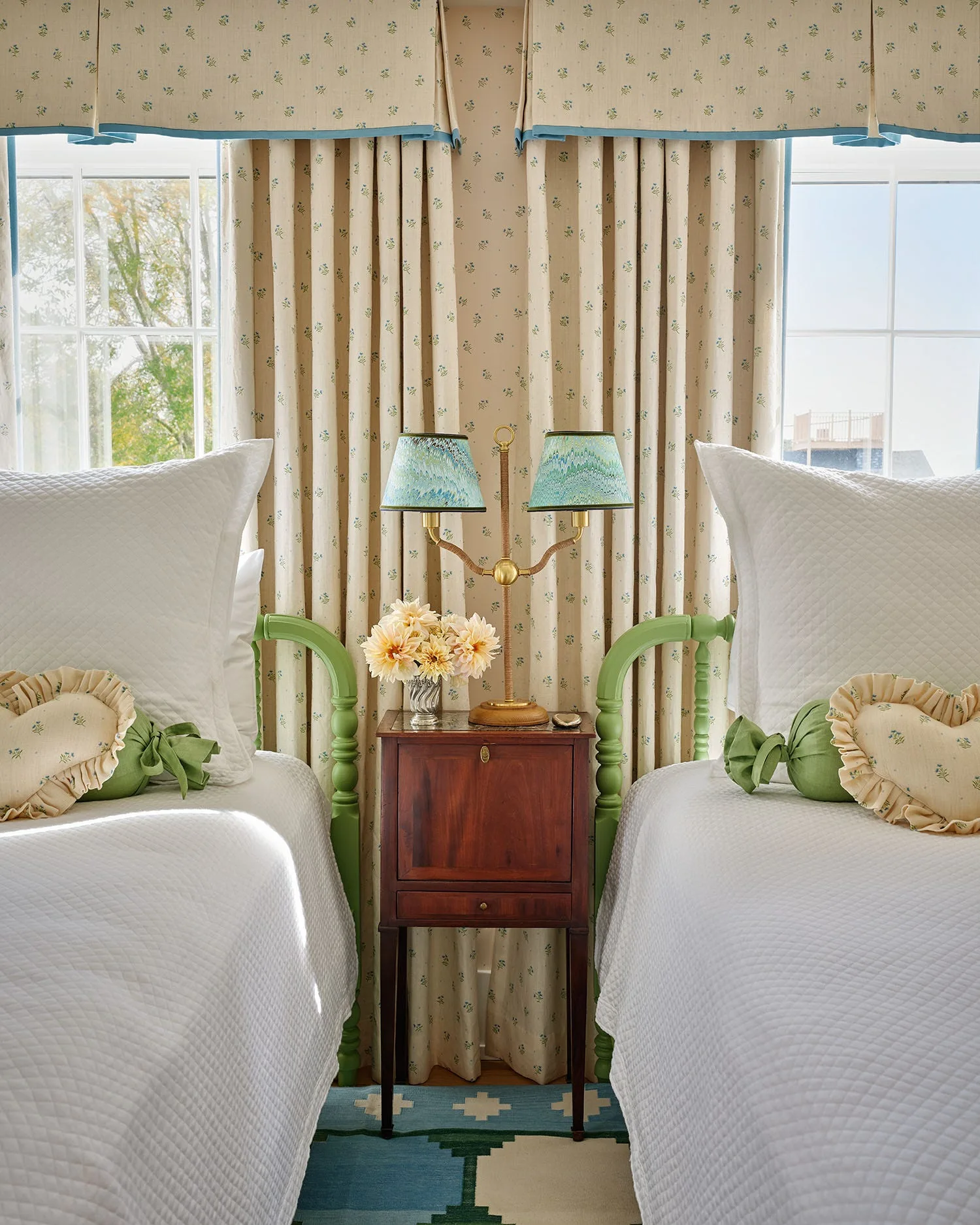 Two twin beds with white quilted bedding and green headboards sit on either side of a wooden nightstand with a brass lamp under floral curtains.