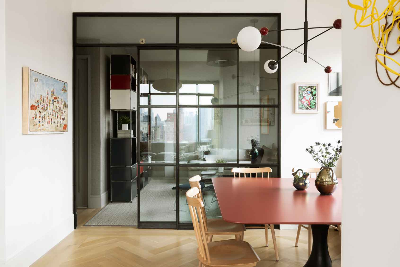 A modern dining room featuring a red table, wooden spindle-back chairs, herringbone wood floors, and a black-framed glass partition wall.