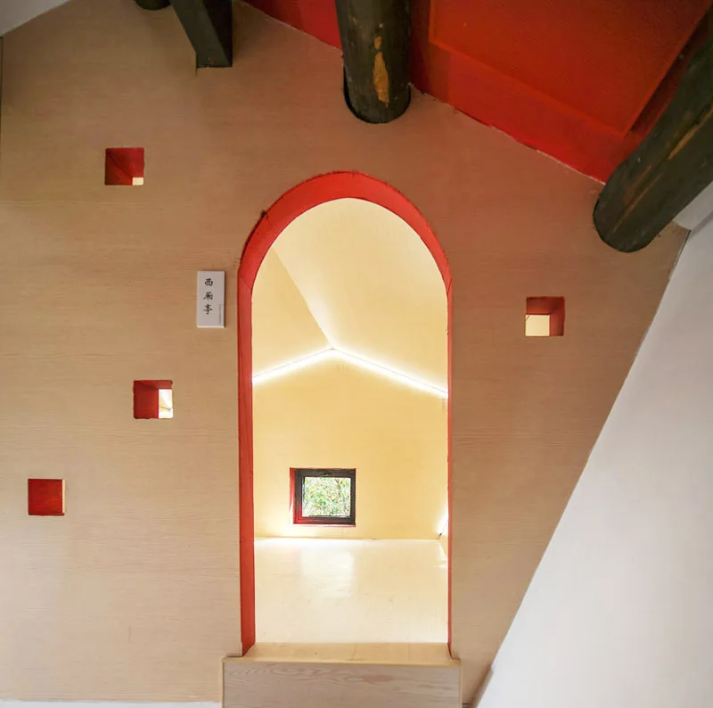 A light wood-paneled wall features small square cutouts and a central archway with red trim leading into a minimalist attic room with linear lighting.