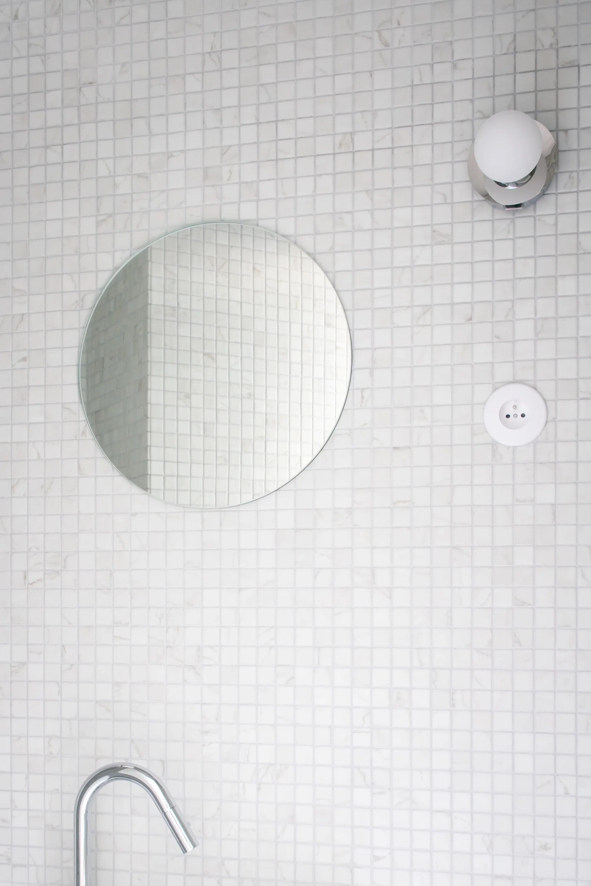 A frameless circular mirror hangs on a white mosaic-tiled wall beside a globe sconce and above a polished chrome faucet.