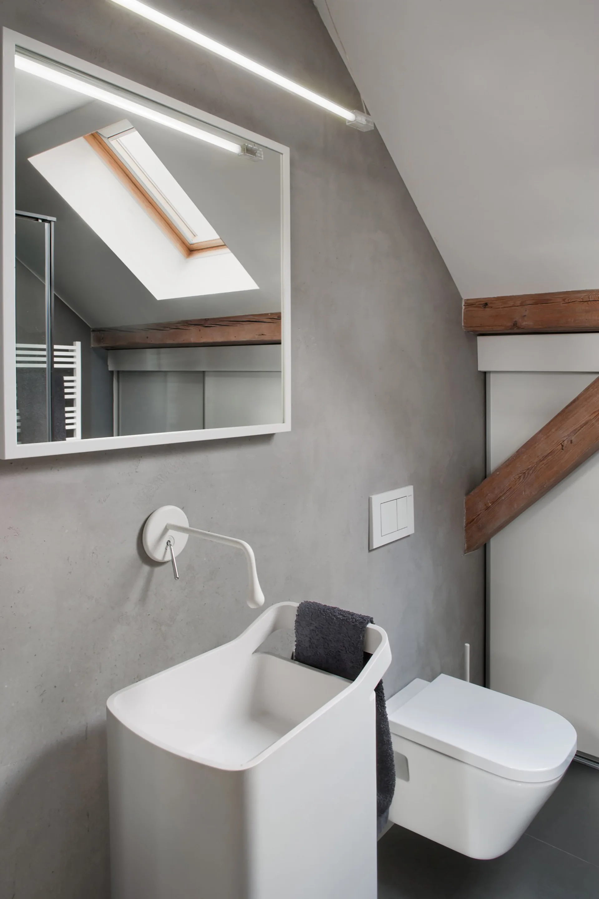 A modern attic bathroom featuring a white wall-mounted sink and toilet set against a grey concrete wall with an exposed wooden beam.