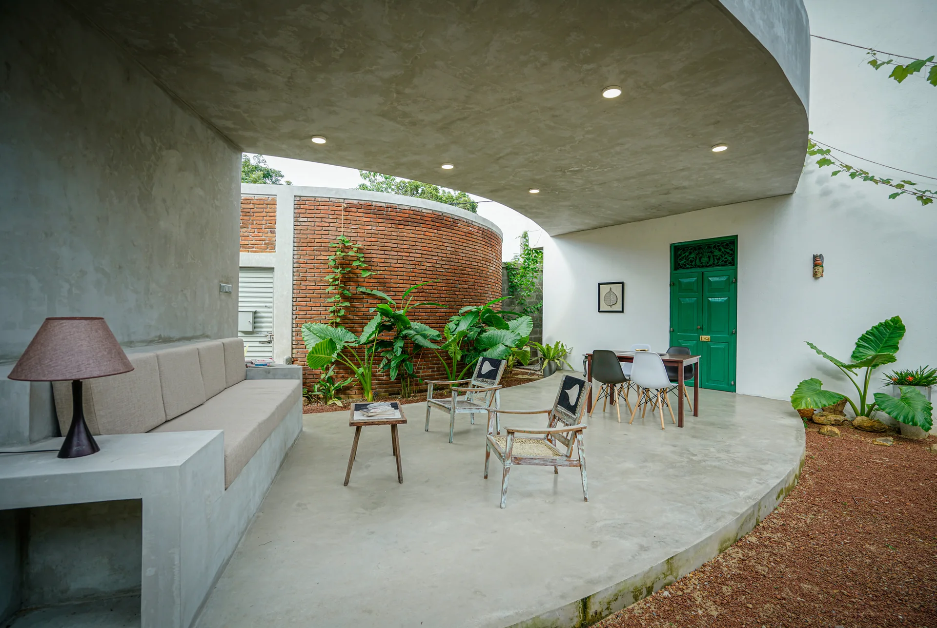 A covered concrete patio features a built-in sofa, rustic wooden armchairs, and a curved red brick wall under a concrete canopy.
