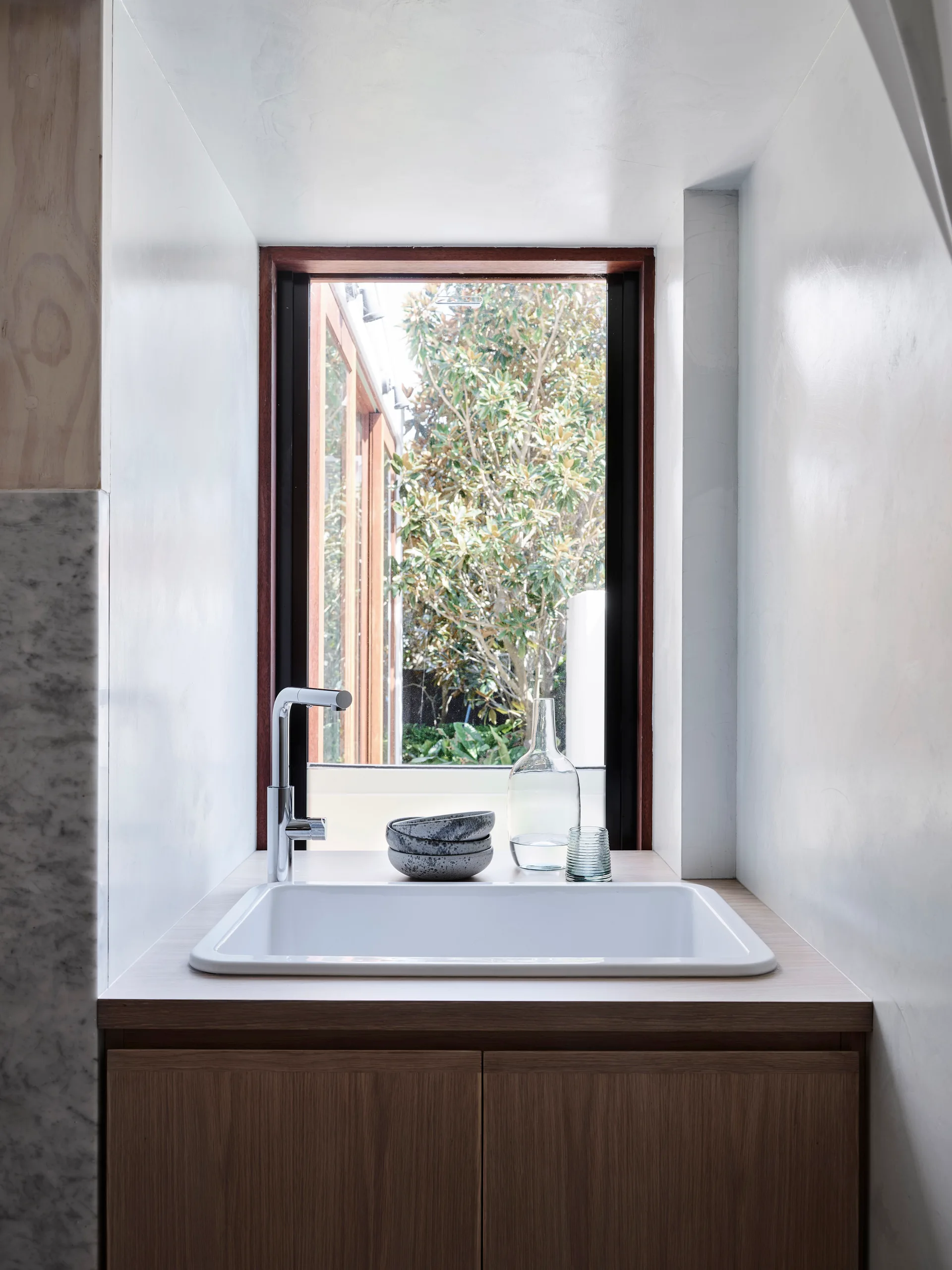 A modern wood vanity features a white rectangular drop-in sink, chrome faucet, and a tall vertical window showing an outdoor garden.