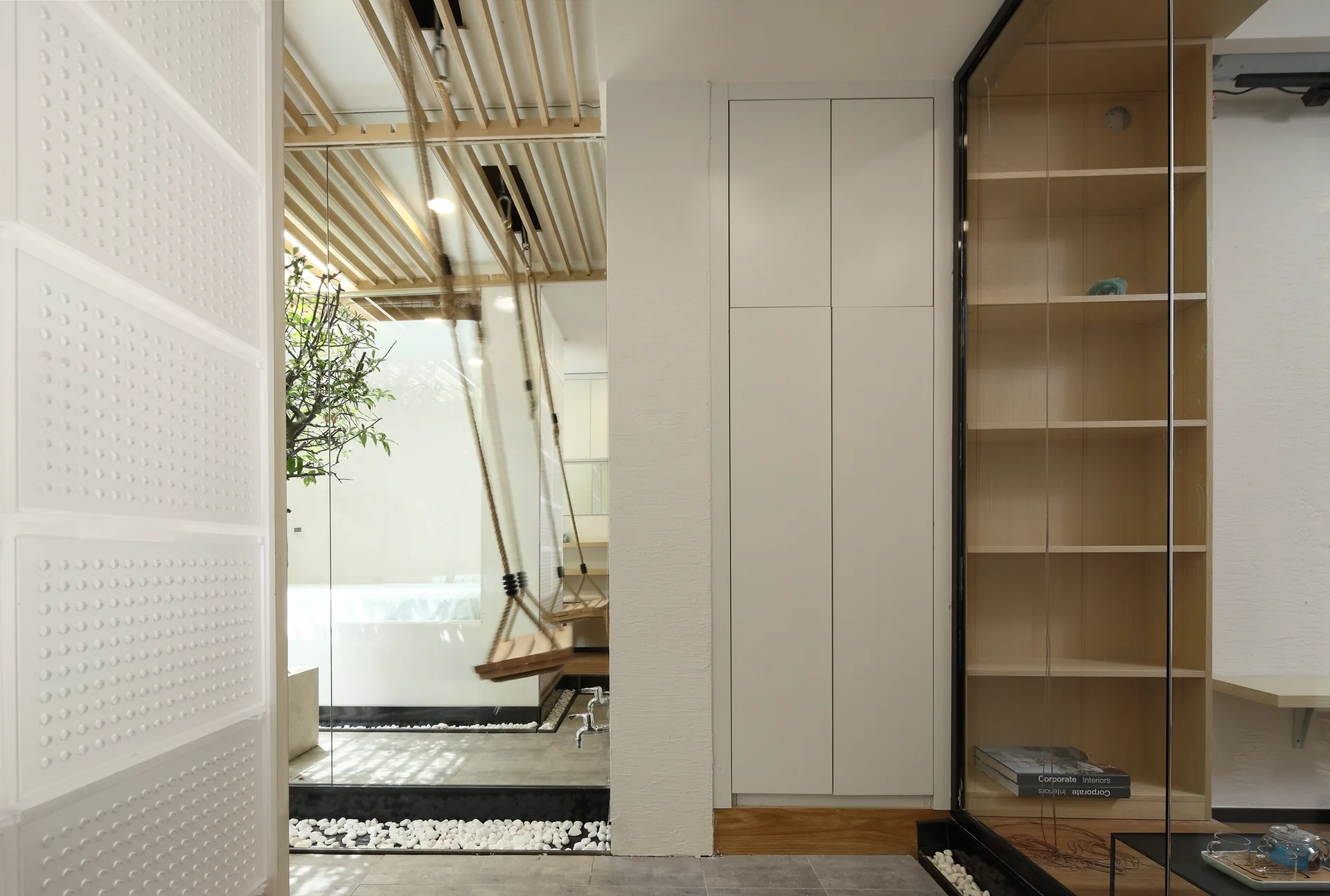 A modern interior space features cream built-in cabinets, a glass display case, and a glass-partitioned area with an indoor wooden swing over white pebbles.