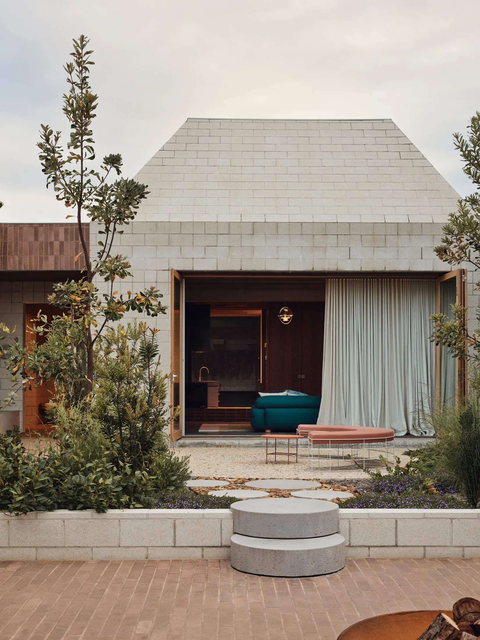 A concrete block house facade features open glass doors revealing a teal sofa inside, with circular concrete steps leading to a landscaped patio.