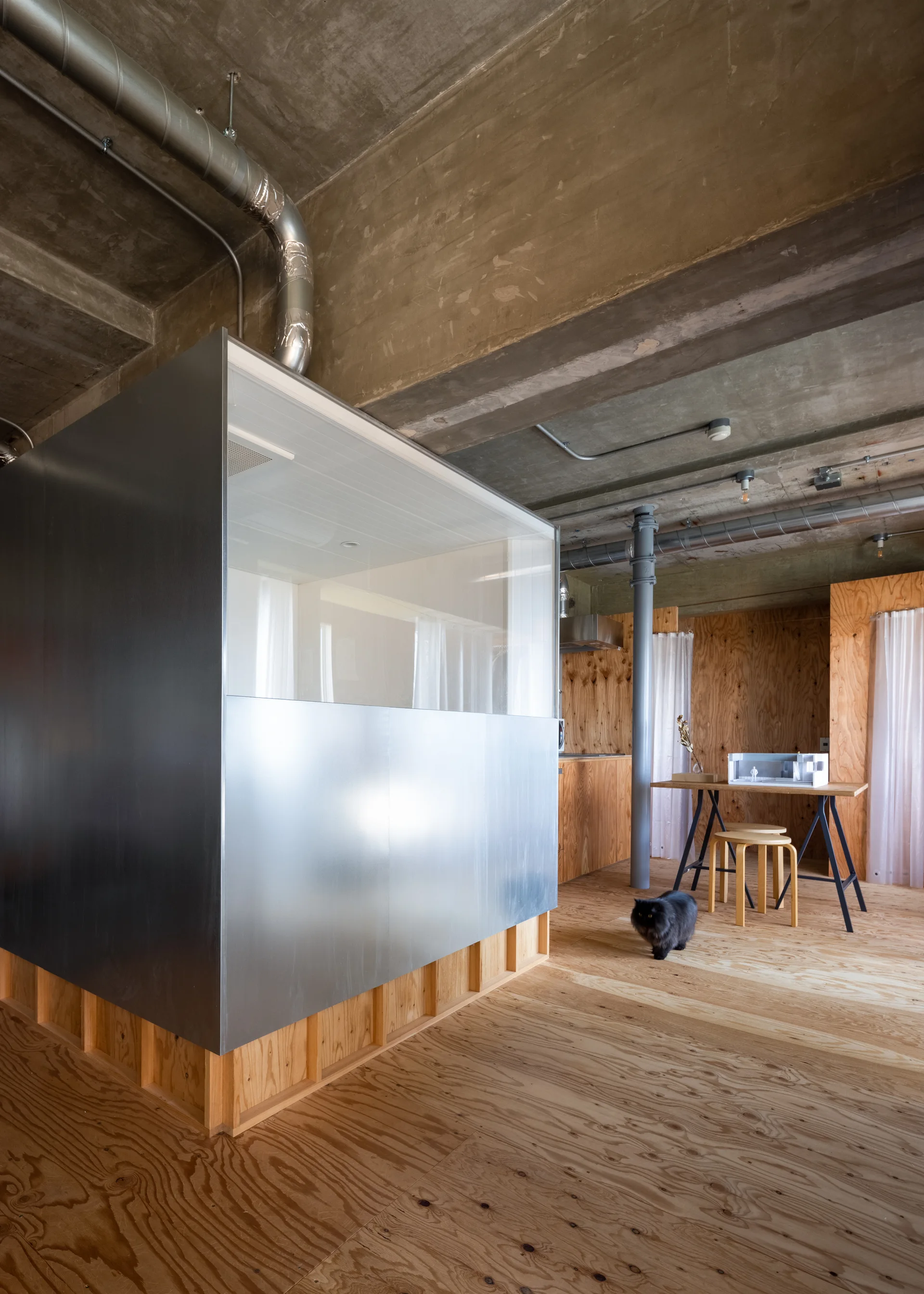 An industrial loft features a large stainless steel modular unit, plywood paneling on walls and floors, and an exposed concrete ceiling with ductwork.