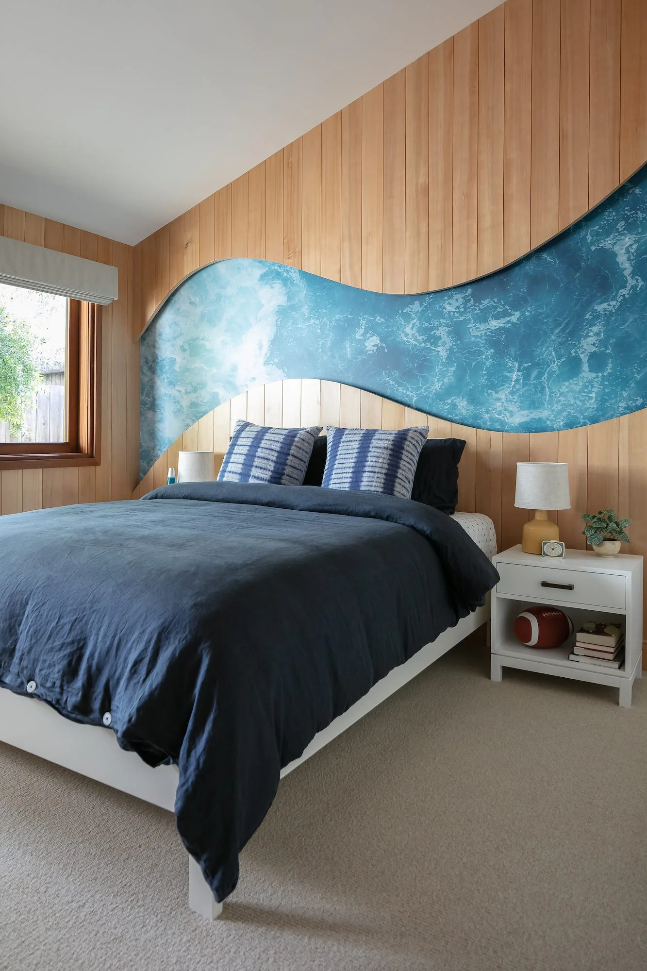 A bedroom features a bed with navy linens, a white nightstand, and a custom accent wall made of vertical wood paneling with a blue wave mural insert.